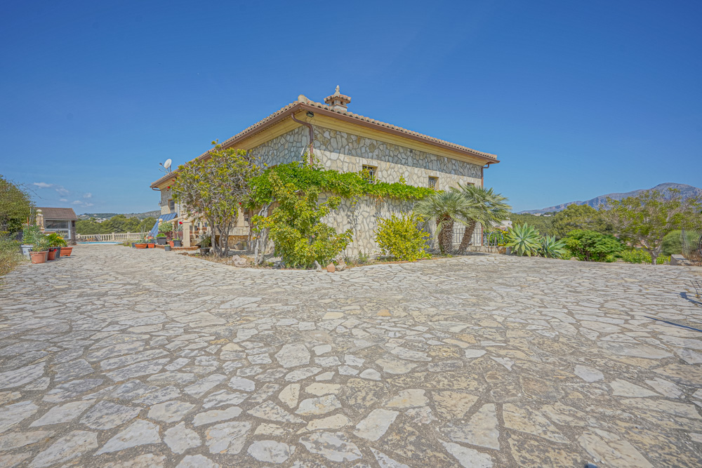 Exterieur van een natuurstenen villa met een ruime geplaveide oprit, mediterrane beplanting met palmbomen en een traditioneel pannendak.