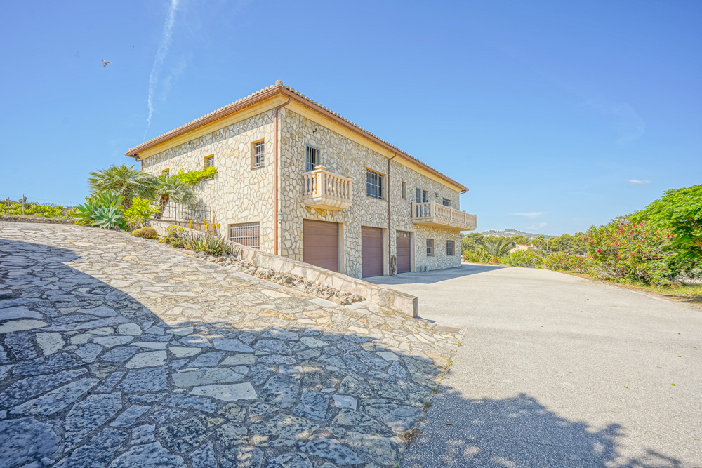 Gevel van natuursteen met een drievoudige garage, geplaveide oprit en klassieke balkons, gelegen in een mediterrane tuin onder een blauwe hemel.