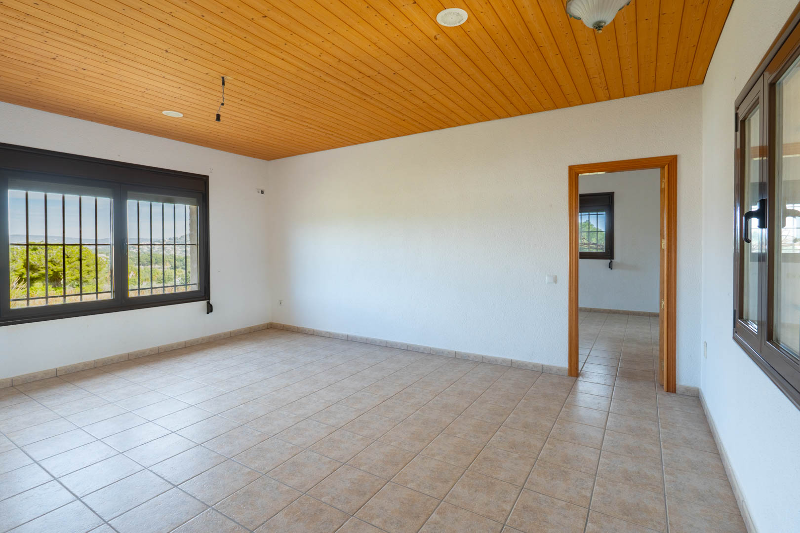 Kamer met houten plafondafwerking, beige tegelvloer en donkere raamkozijnen met veiligheidsstangen en uitzicht op de groene omgeving.