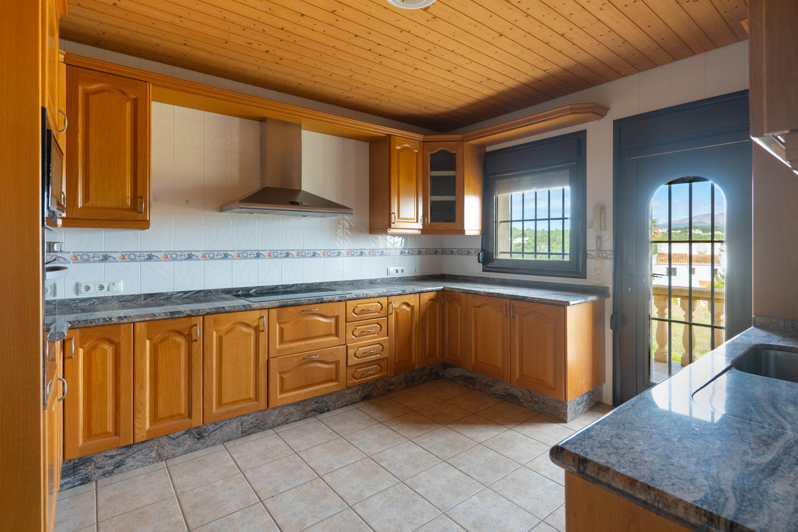 Keuken met houten kasten, granieten werkbladen en een houten plafond. Voorzien van een RVS afzuigkap en toegang tot een terras met uitzicht op de omgeving.