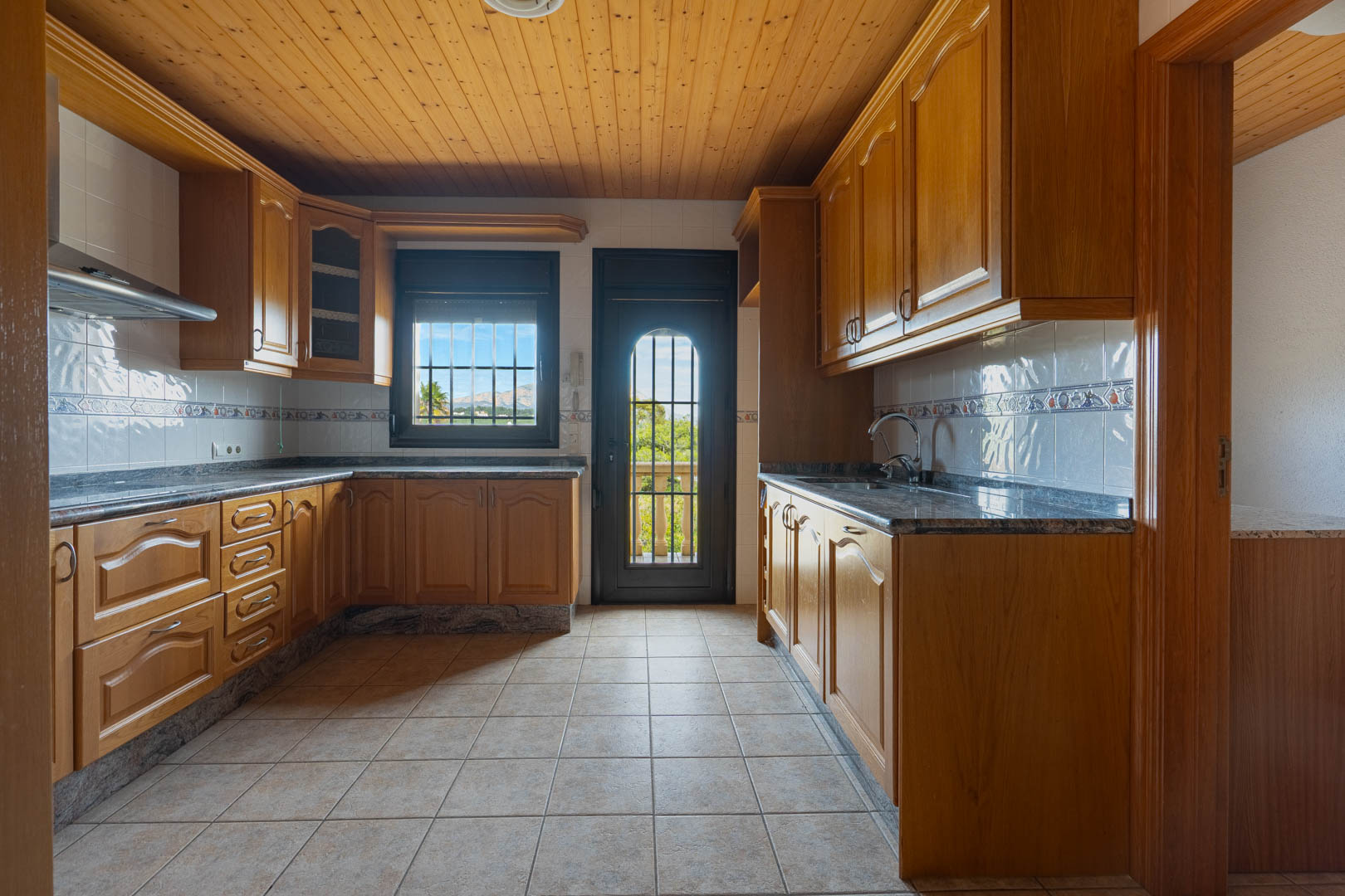 Keuken voorzien van houten kasten, granieten werkbladen en een houten plafond. De ruimte heeft een tegelvloer en een deur naar een balkon met uitzicht op de bergen.