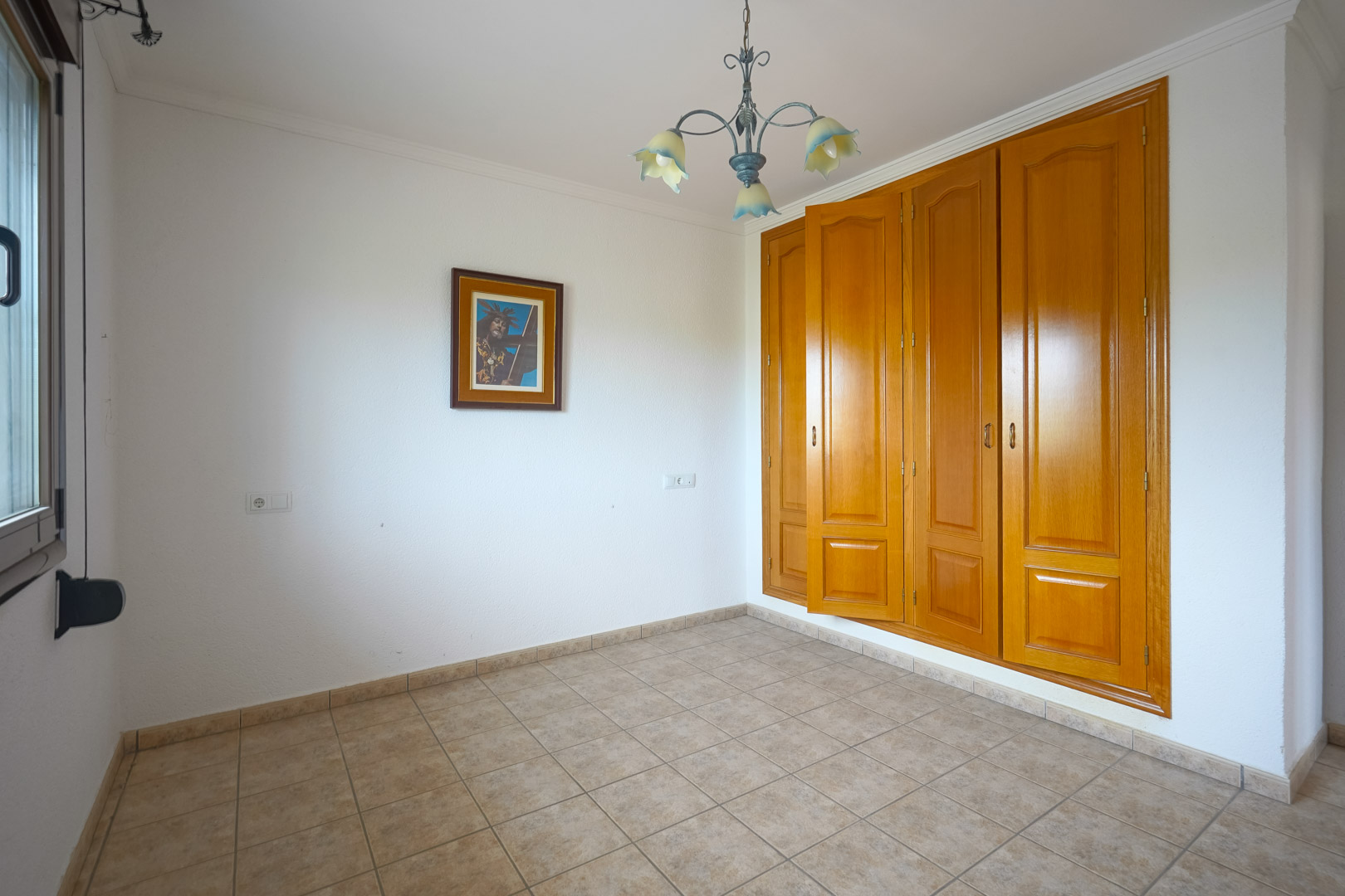 Lege kamer met een grote ingebouwde houten kledingkast met vier deuren, beige keramische tegelvloer en een decoratieve kroonluchter met bloemvormige kappen.