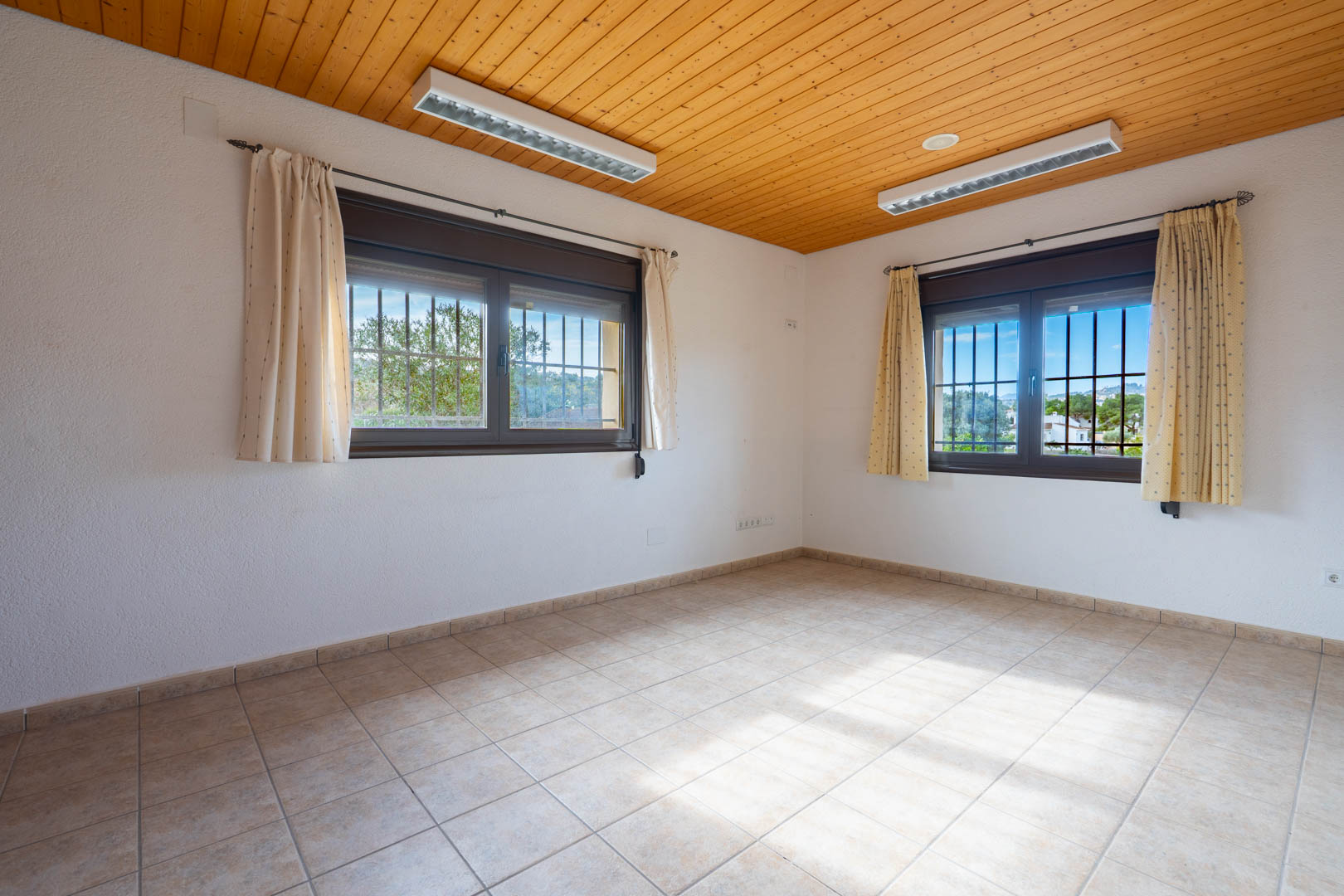 Lege kamer met een plafond van houten schroten, een keramische tegelvloer en twee ramen voorzien van beveiligingstralies en gordijnen.