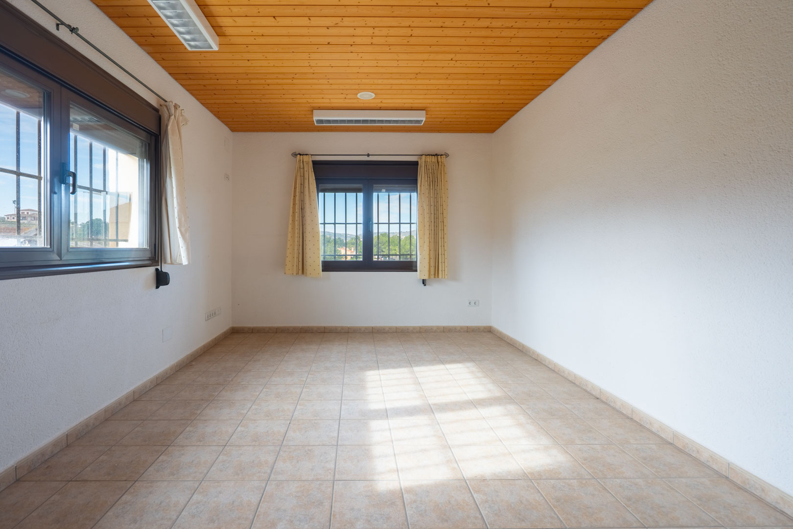 Lichte lege kamer met een houten plafond, beige tegelvloer en twee ramen met beveiligingstralies en crèmekleurige gordijnen.