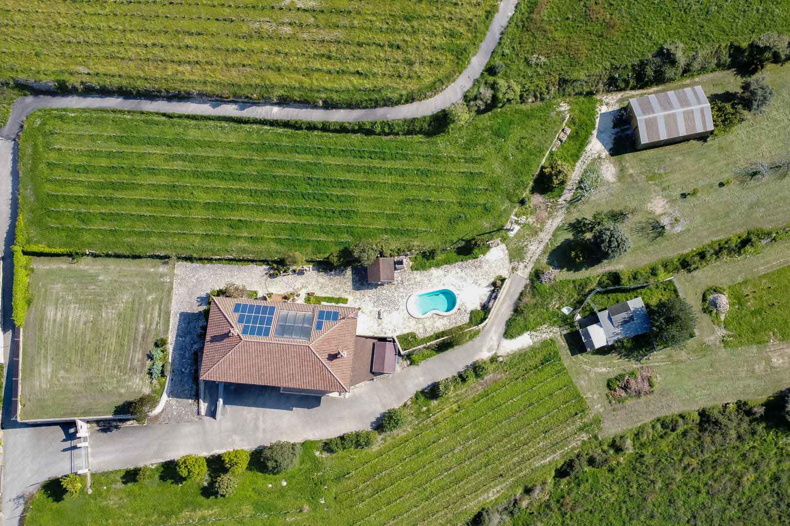 Luchtfoto van een landelijk perceel met zonnepanelen op het dak, een niervormig zwembad en uitgestrekte landbouwgronden met strakke plantrijen.