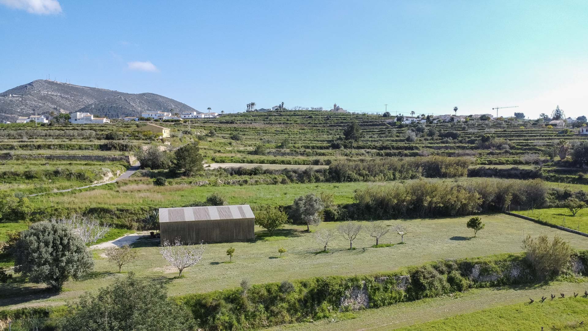 Luchtfoto van terrasvormige heuvels met traditionele stenen muren, een houten schuur en jonge fruitbomen in een landelijk landschap.
