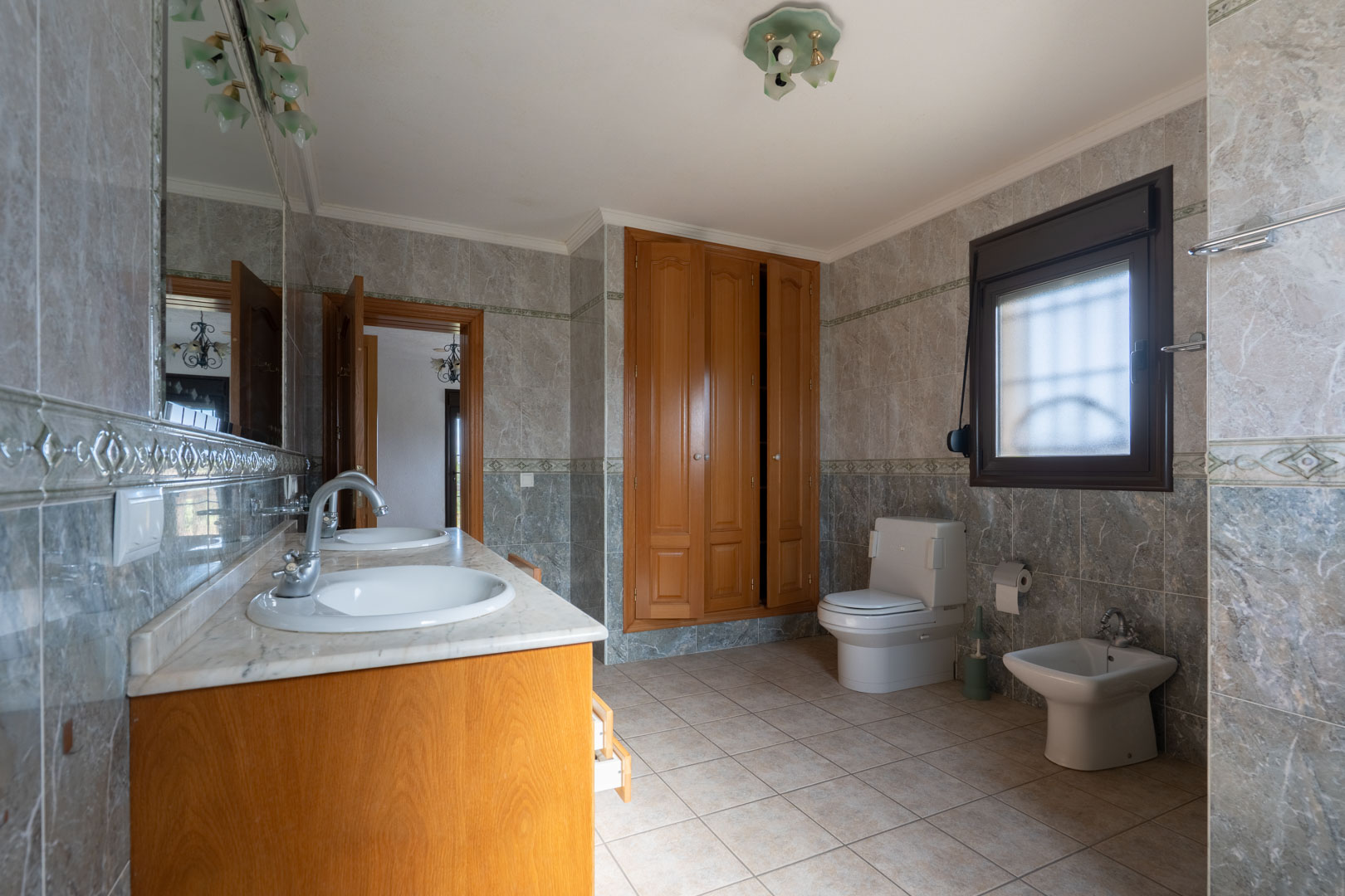 Ruime badkamer met dubbele marmeren wastafel, houten inbouwkasten en volledig betegelde wanden met decoratieve sierlijsten. Inclusief toilet en bidet.