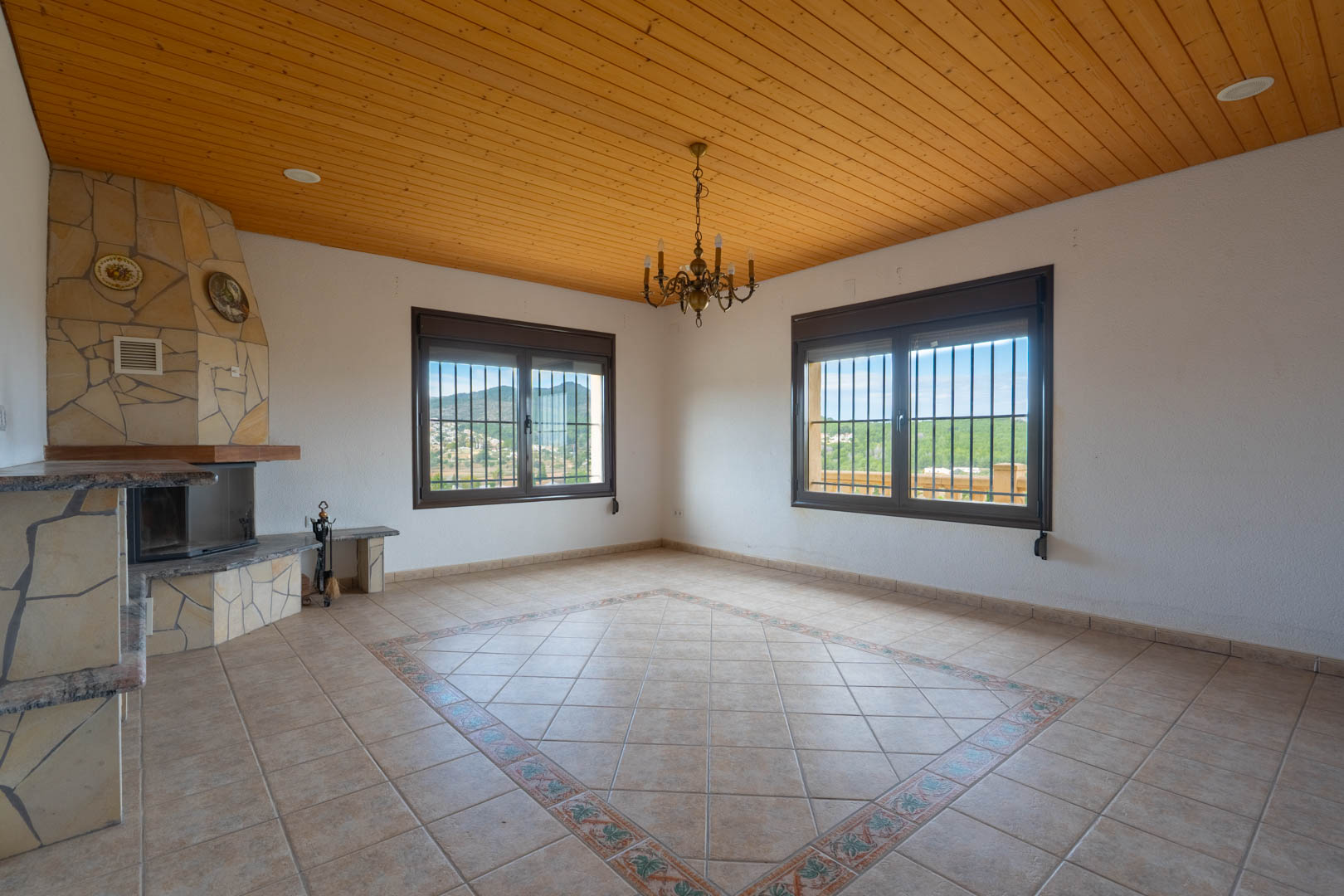 Woonkamer met houten plafond, een hoekhaard bekleed met natuursteen en een tegelvloer met decoratieve rand. Grote ramen kijken uit op het heuvelachtige landschap.