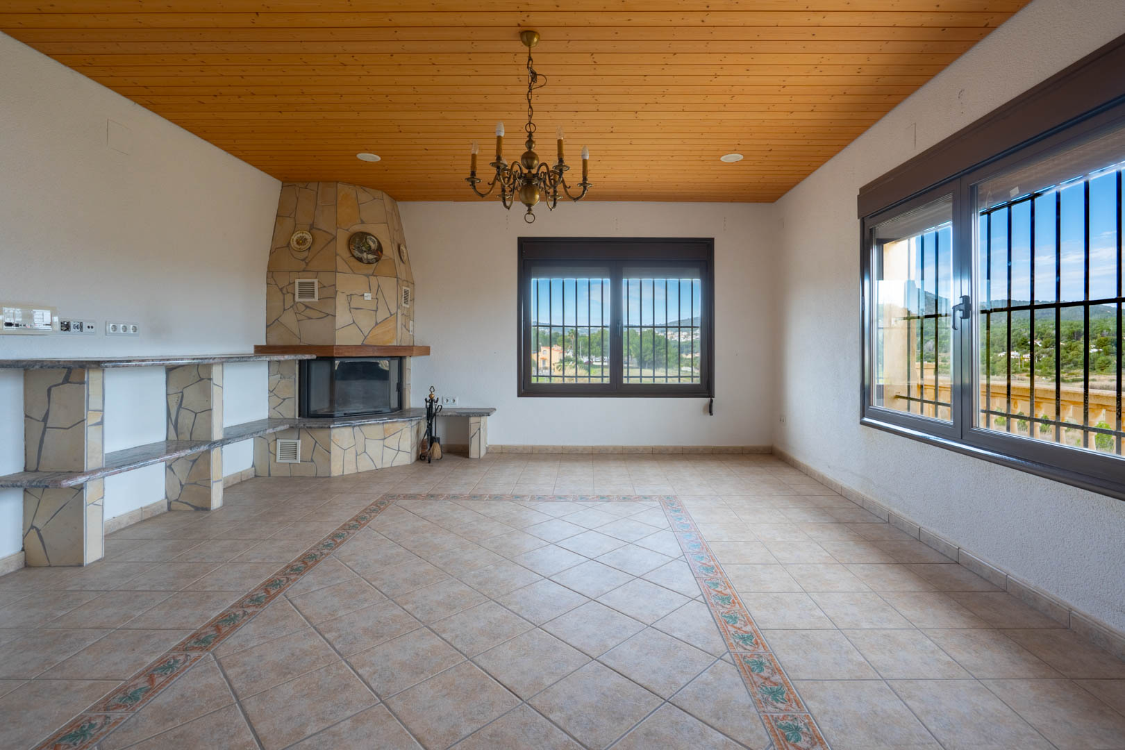 Woonkamer met houten plafond, natuurstenen open haard en tegelvloer met decoratieve rand. Grote ramen bieden uitzicht op de landelijke omgeving.