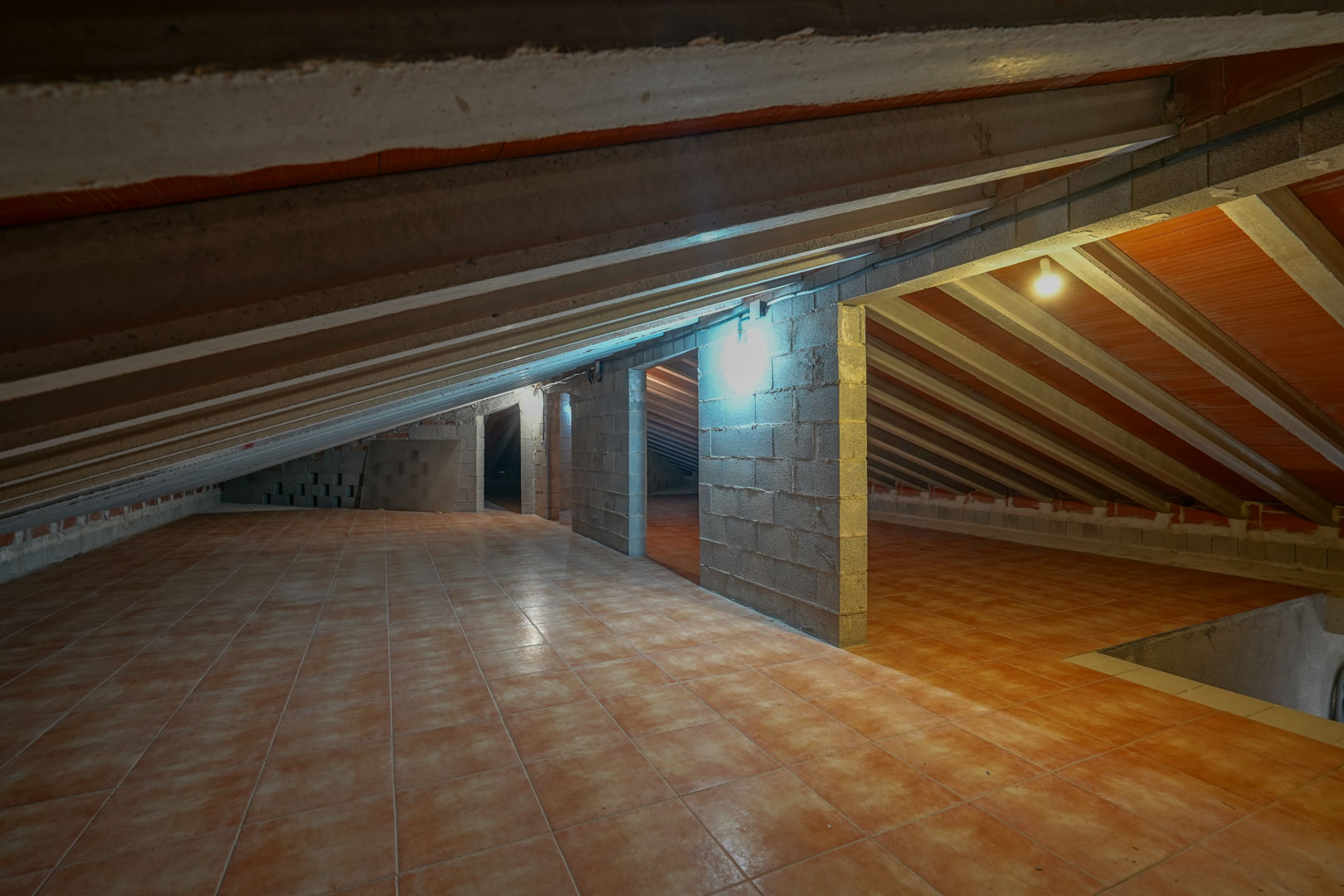 Zolderberging met terracotta vloertegels, zichtbare betonnen plafondbalken en scheidingswanden van betonblokken met verlichting.