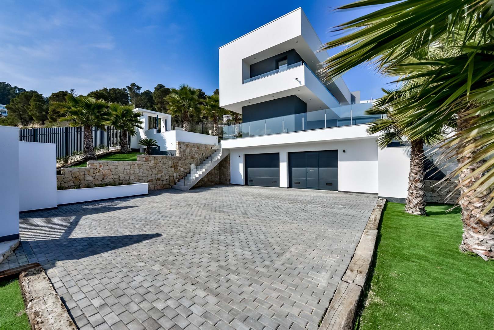Eigentijdse gevel met dubbele garage, natuurstenen muren en een geplaveide oprit omgeven door mediterrane beplanting.