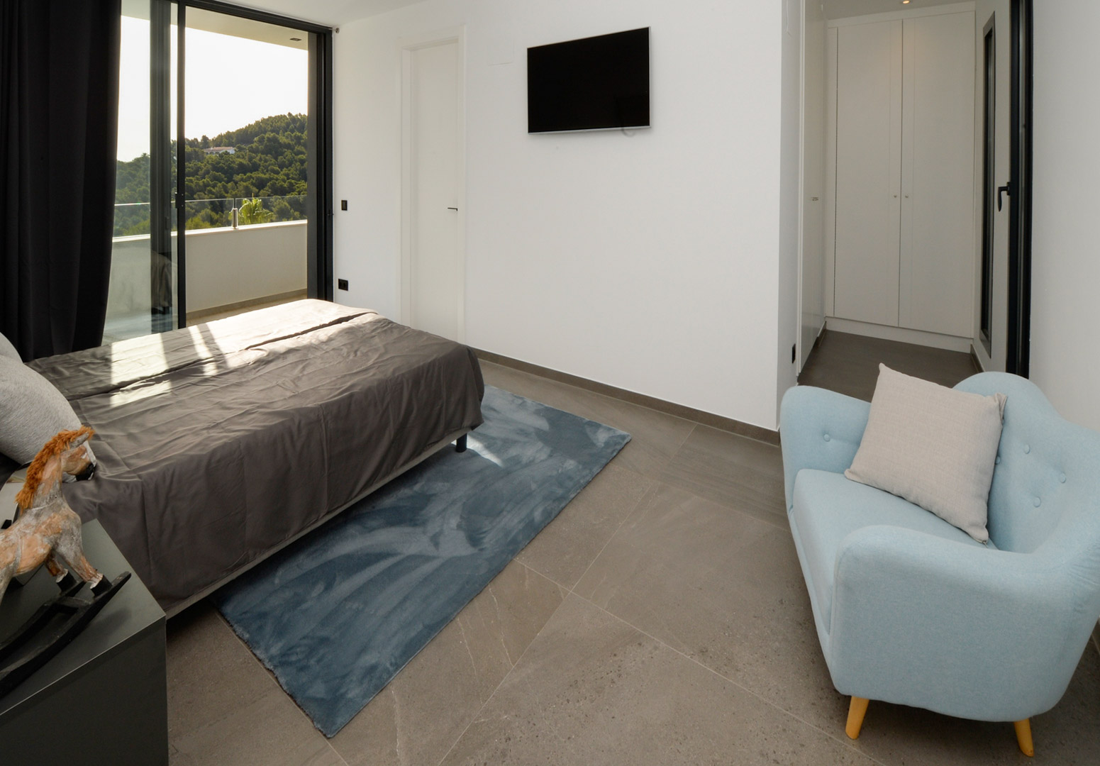 Eigentijdse slaapkamer met toegang tot het terras, grijze tegelvloer, witte inbouwkasten en panoramisch uitzicht over de groene heuvels.