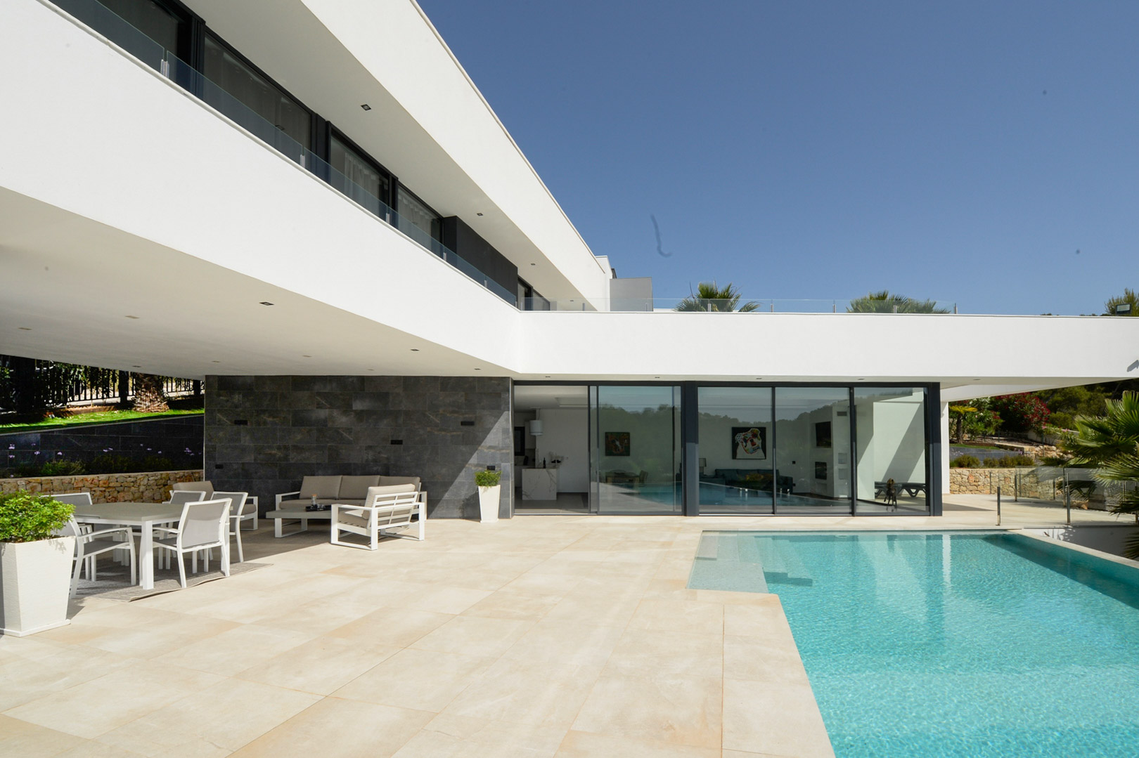Hoofdterras met infinity zwembad, overdekte eethoek en minimalistische architectuur met kamerhoge glazen schuifpuien en natuurstenen wandbekleding.