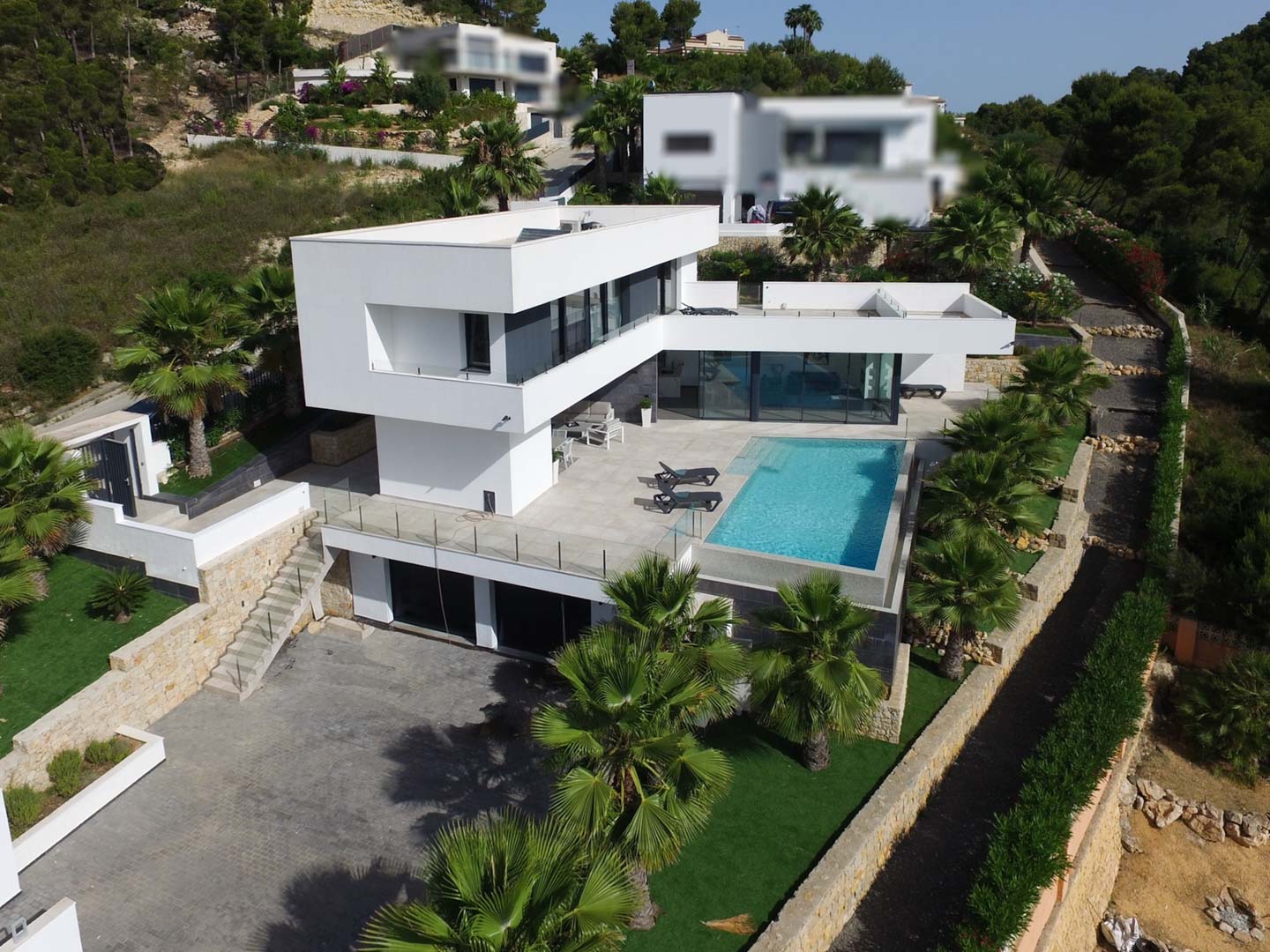 Luchtfoto van de moderne villa met witte architectuur, infinity zwembad, stenen terrassen en een mediterrane tuin met palmbomen.