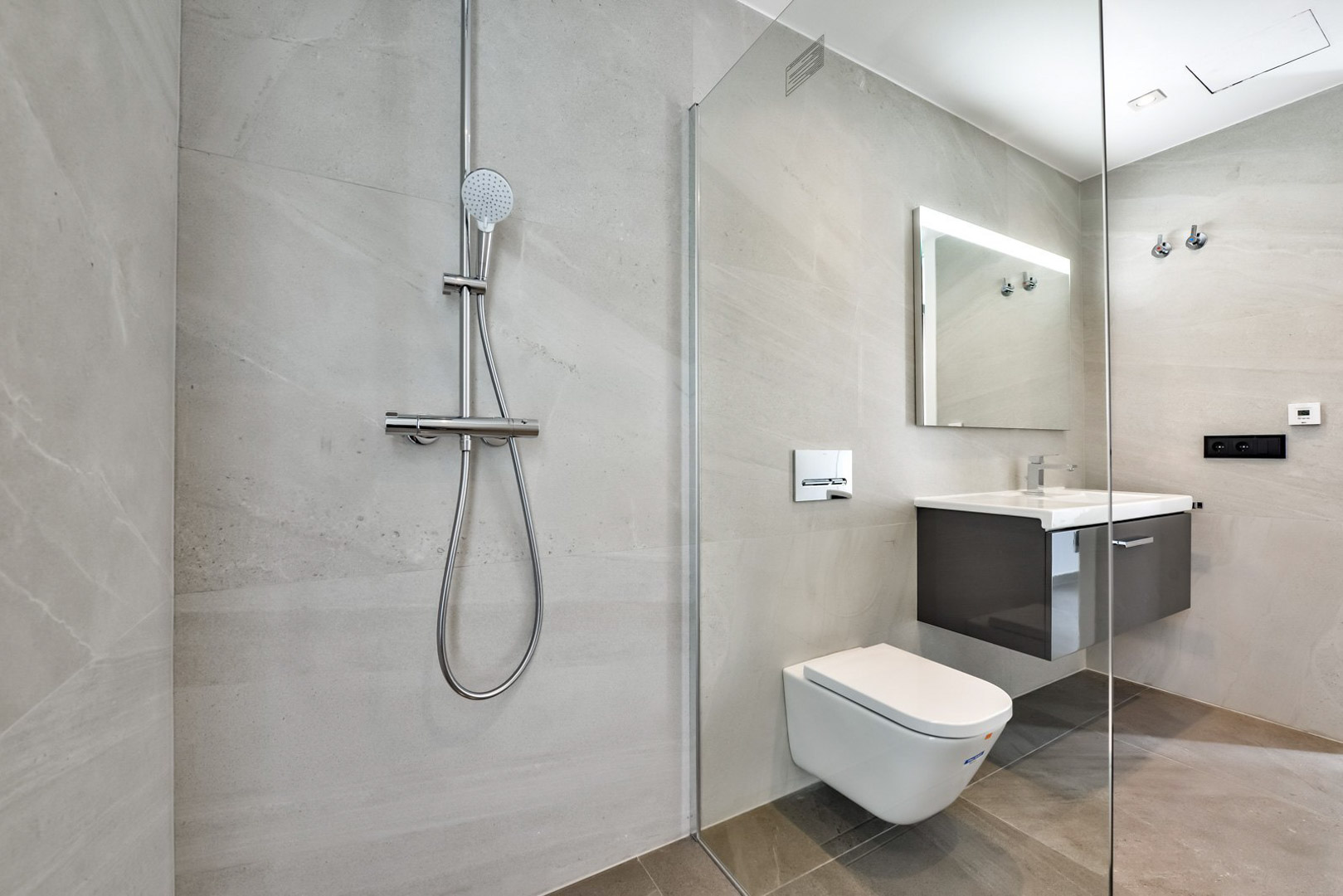 Moderne badkamer met inloopdouche, glazen wand, hangend toilet en zwevend wastafelmeubel. Grootformaat grijze tegels en een verlichte spiegel zorgen voor een strakke afwerking.