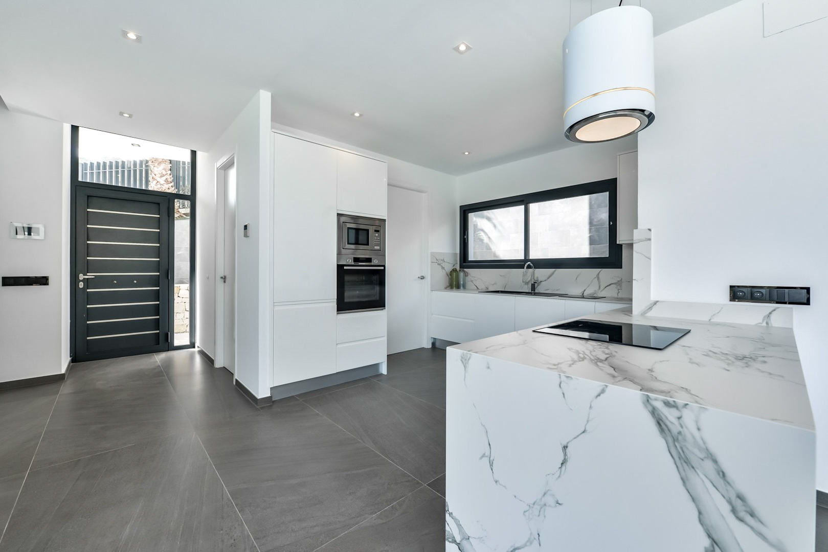 Moderne designkeuken met kookeiland in marmerlook, inbouwapparatuur en een cilindrische afzuigkap naast de zwarte minimalistische voordeur.