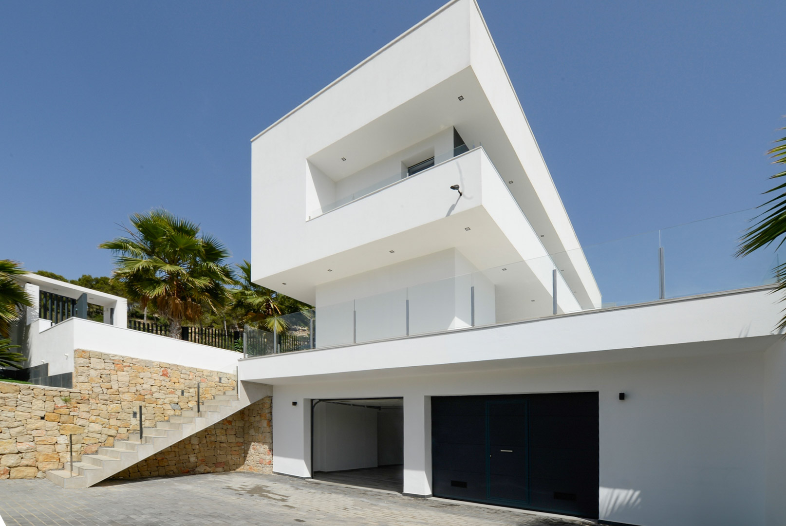 Moderne gevel met witte kubistische vormen, dubbele garage, natuurstenen muren en buitentrap met glazen balustrades.