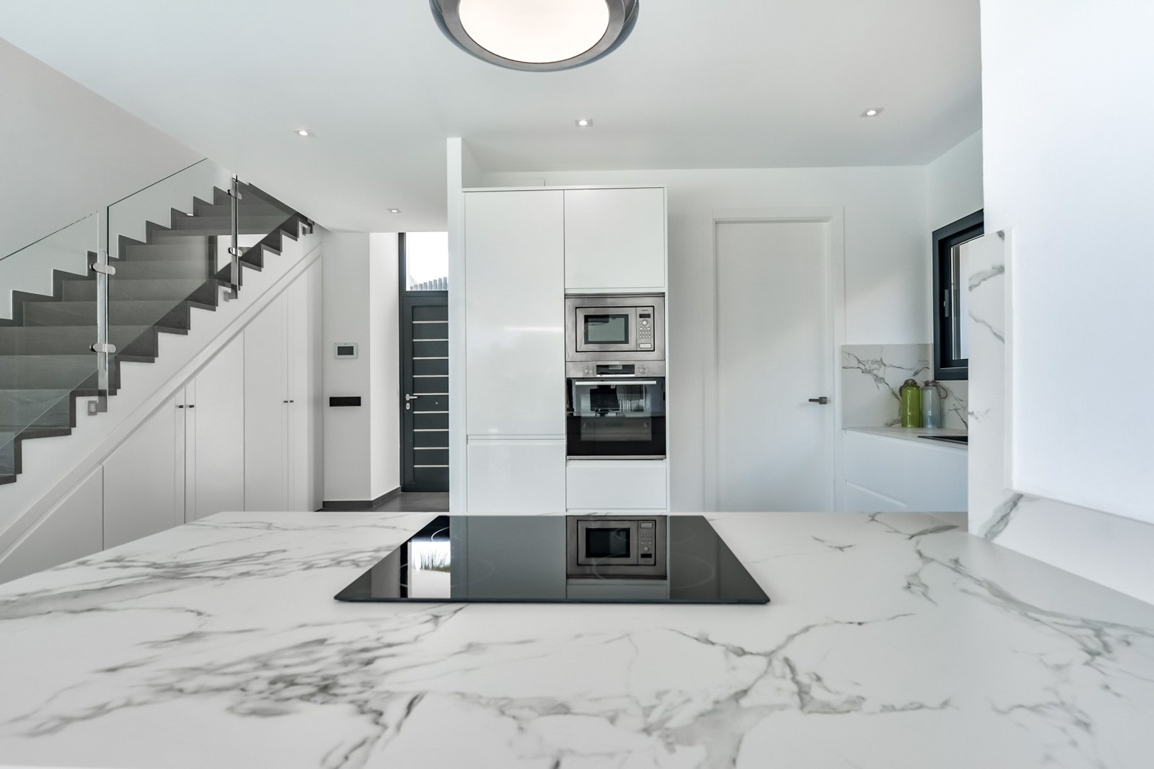 Moderne keuken met marmeren werkblad en inductiekookplaat, grenzend aan een eigentijdse trap met glazen balustrade en ingebouwde kastenwand.