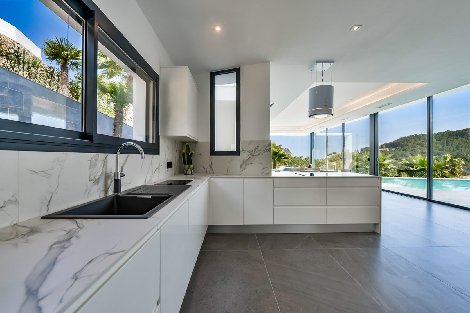 Moderne keuken met marmerlook werkbladen en een cilindrische afzuigkap. Grote glazen puien bieden een direct uitzicht op het overloopzwembad en de omliggende natuur.