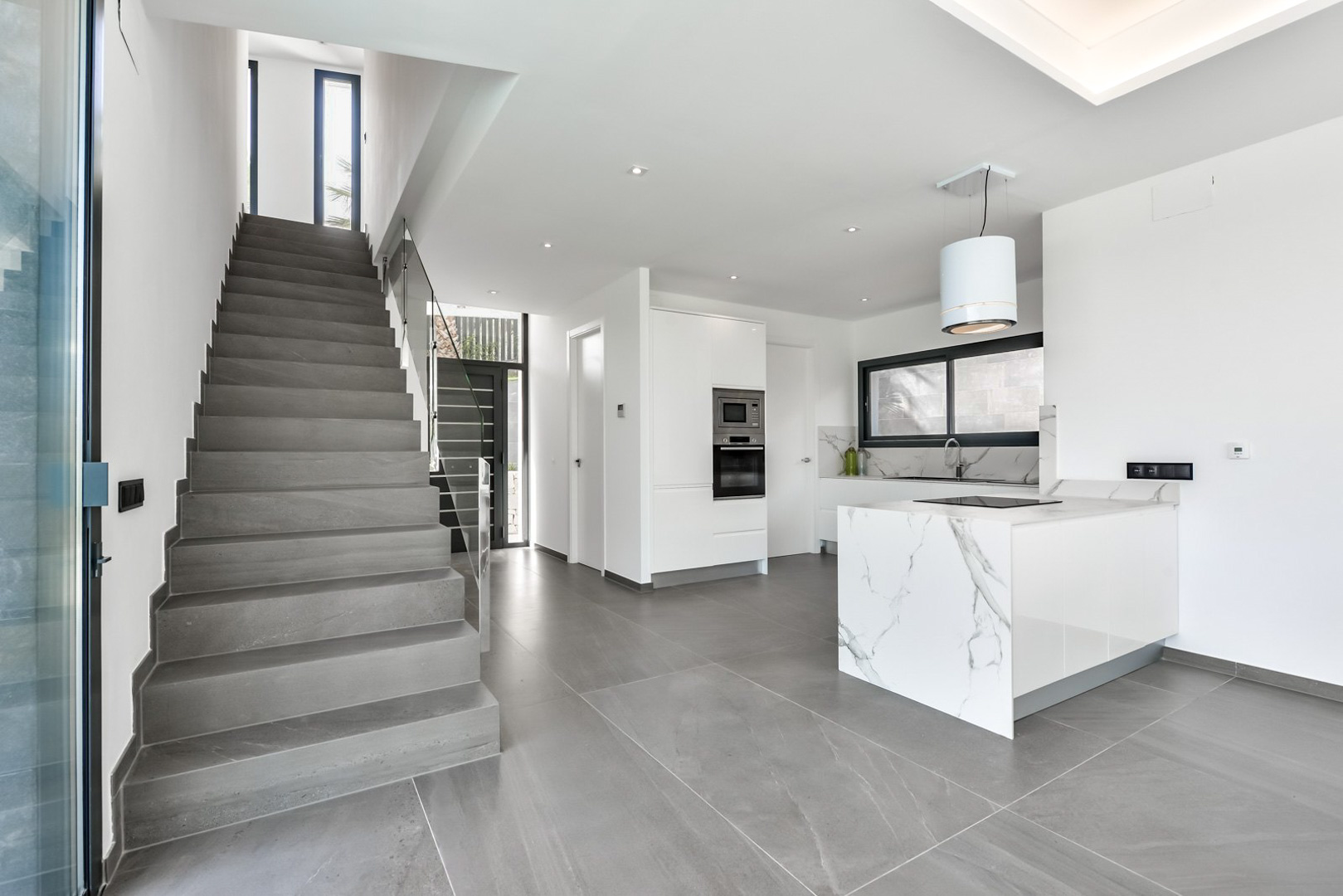 Moderne open keuken met een marmeren kookeiland en inbouwapparatuur, grenzend aan een grijze natuurstenen trap met glazen balustrade.