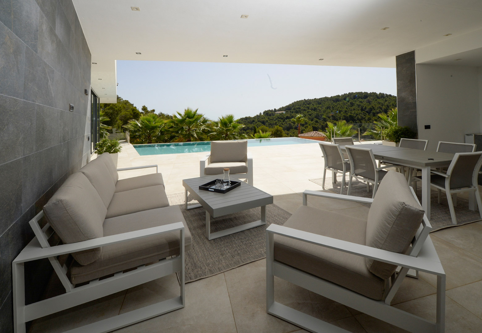Overdekt terras met moderne lounge- en eethoek, uitkijkend over het overloopzwembad en de beboste heuvels van Jávea.