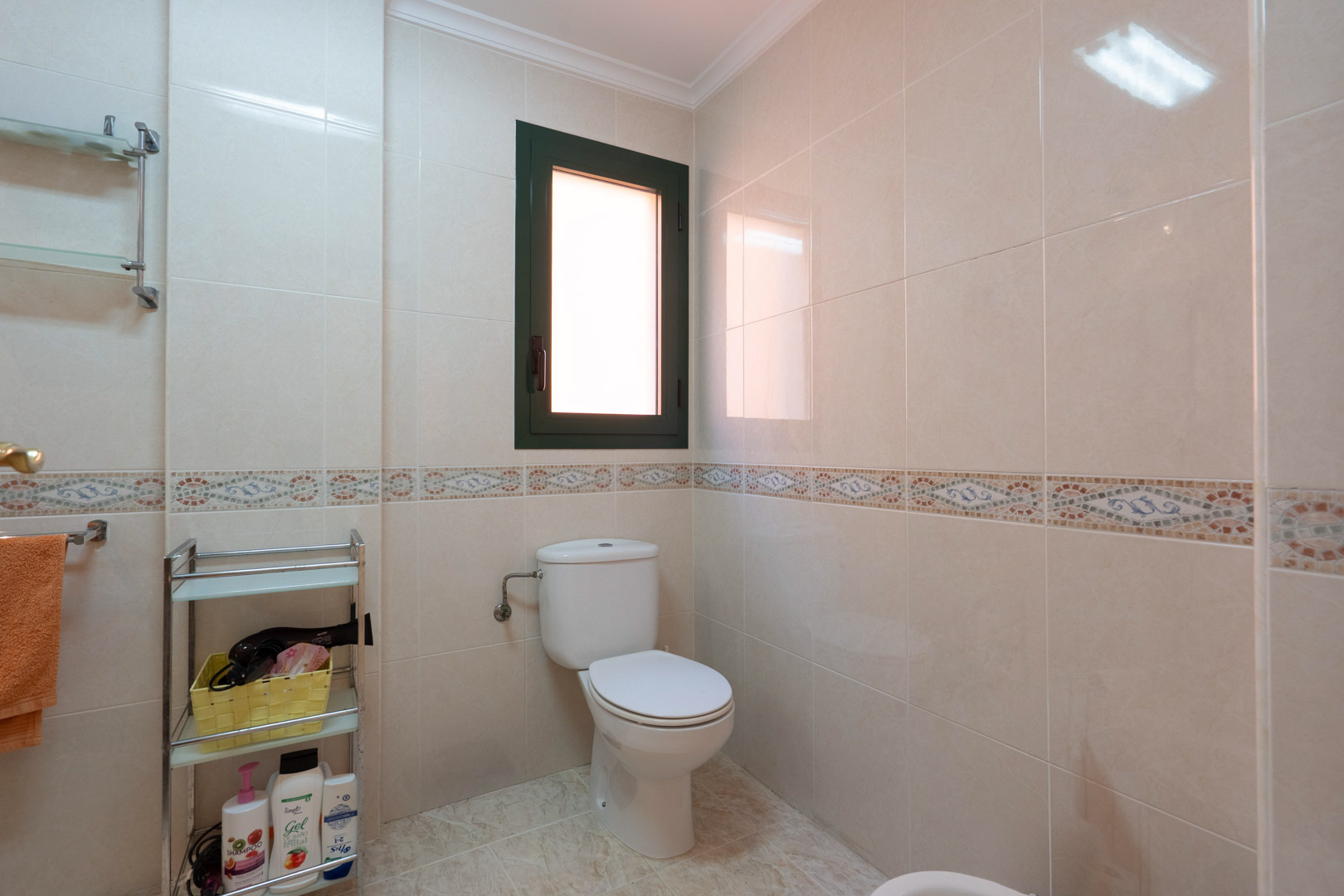 Badkamer met crème wandtegels, een decoratieve mozaïekrand, wit toilet en een raam voor natuurlijke lichtinval en ventilatie.