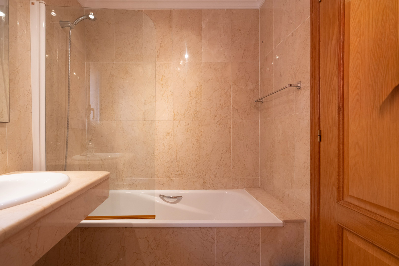 Badkamer met ligbad, glazen douchescherm en wanden van crème marmer. Voorzien van een natuurstenen wastafelblad en een massief houten deur.