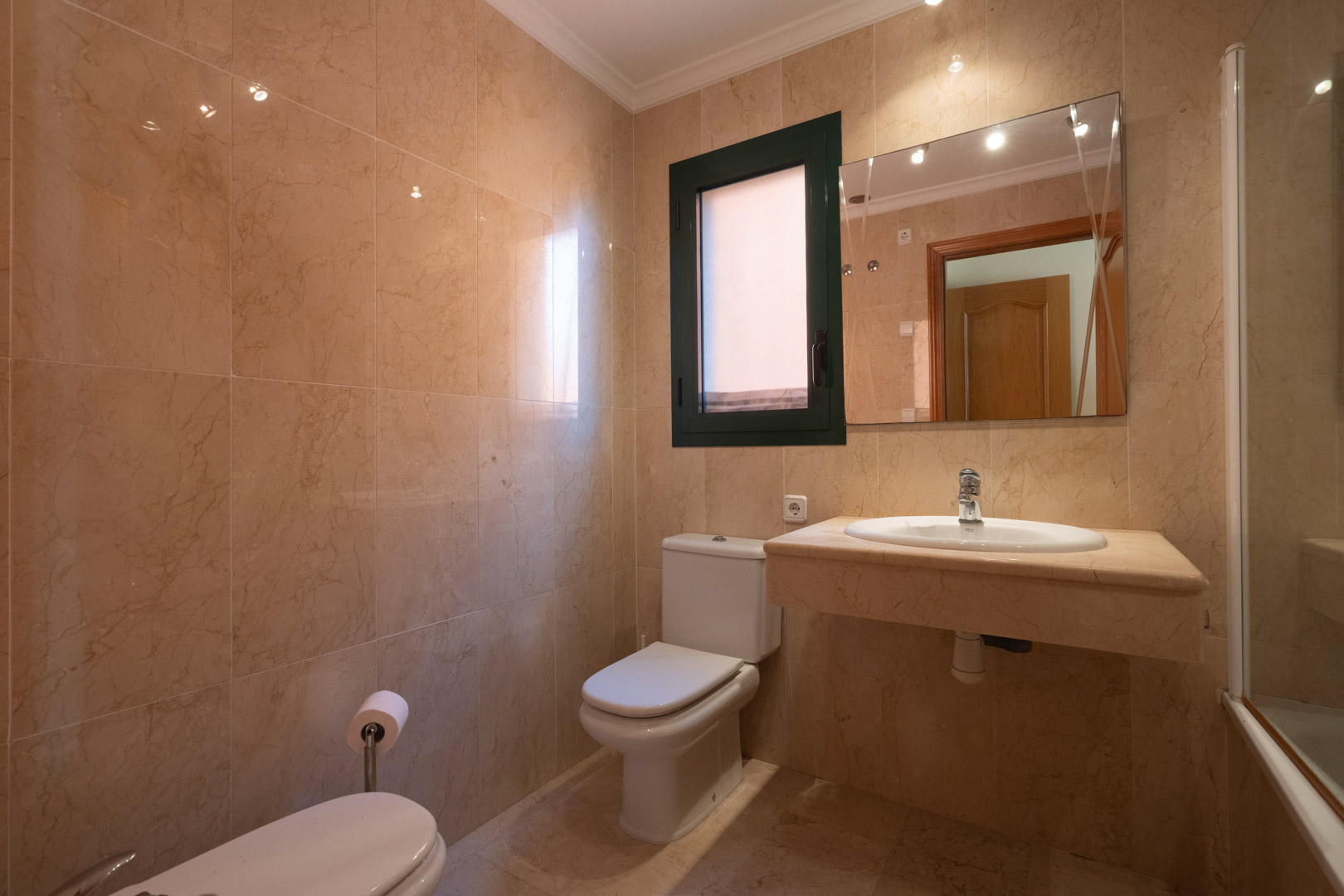 Badkamer volledig afgewerkt met crème marmer, voorzien van een natuurstenen wastafelmeubel, wit sanitair en een raam voor natuurlijke ventilatie.
