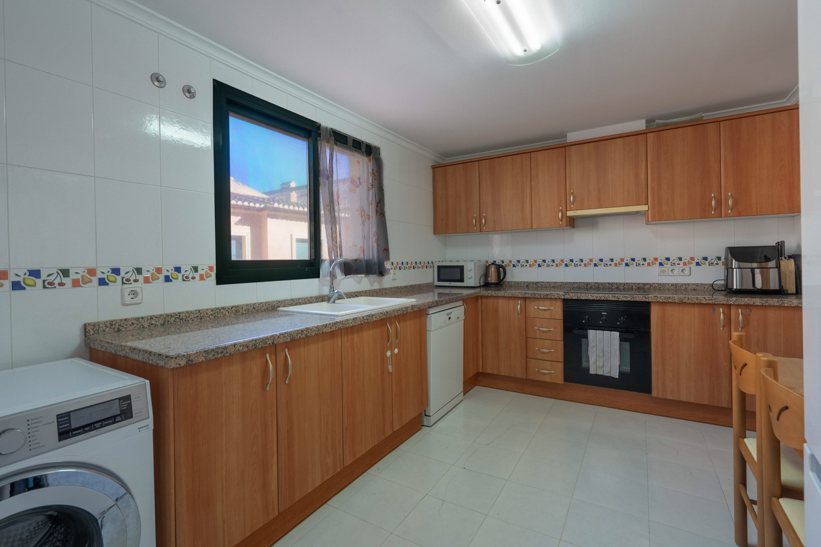 Functionele keuken met houten kasten, granieten werkbladen, decoratieve tegelranden en volledige uitrusting inclusief wasmachine en vaatwasser.