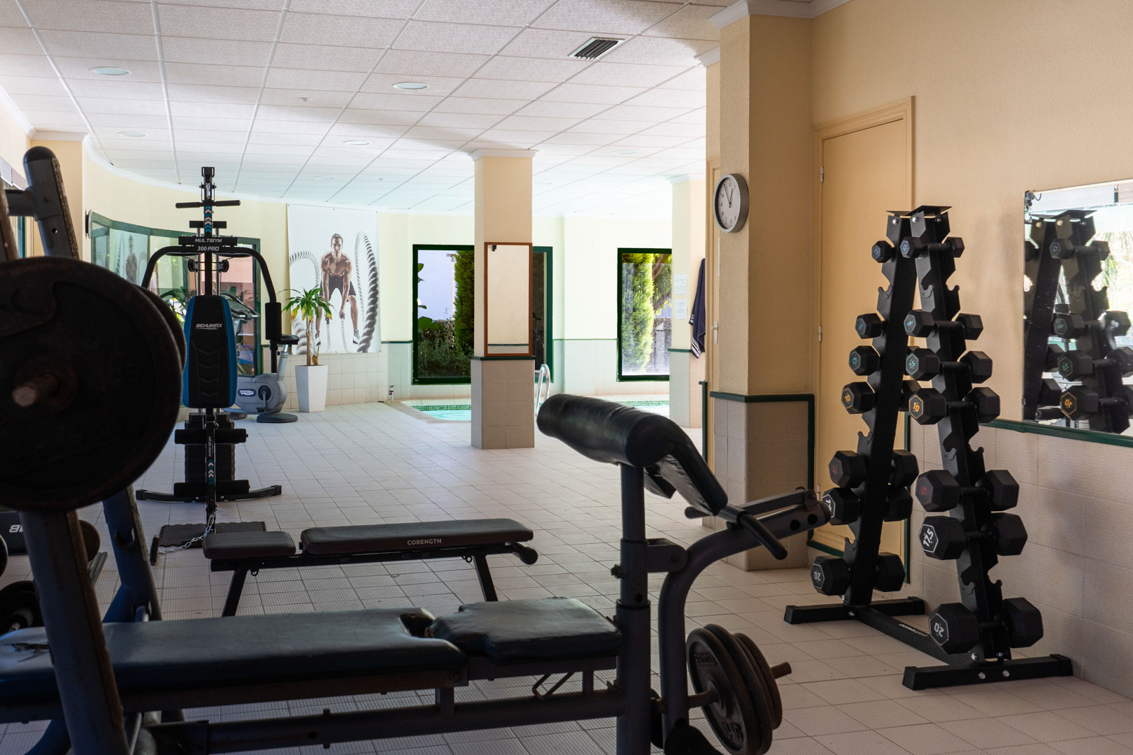Gemeenschappelijke fitnessruimte met krachttoestellen en gewichten, gelegen naast het binnenzwembad met uitzicht op de omliggende tuin.