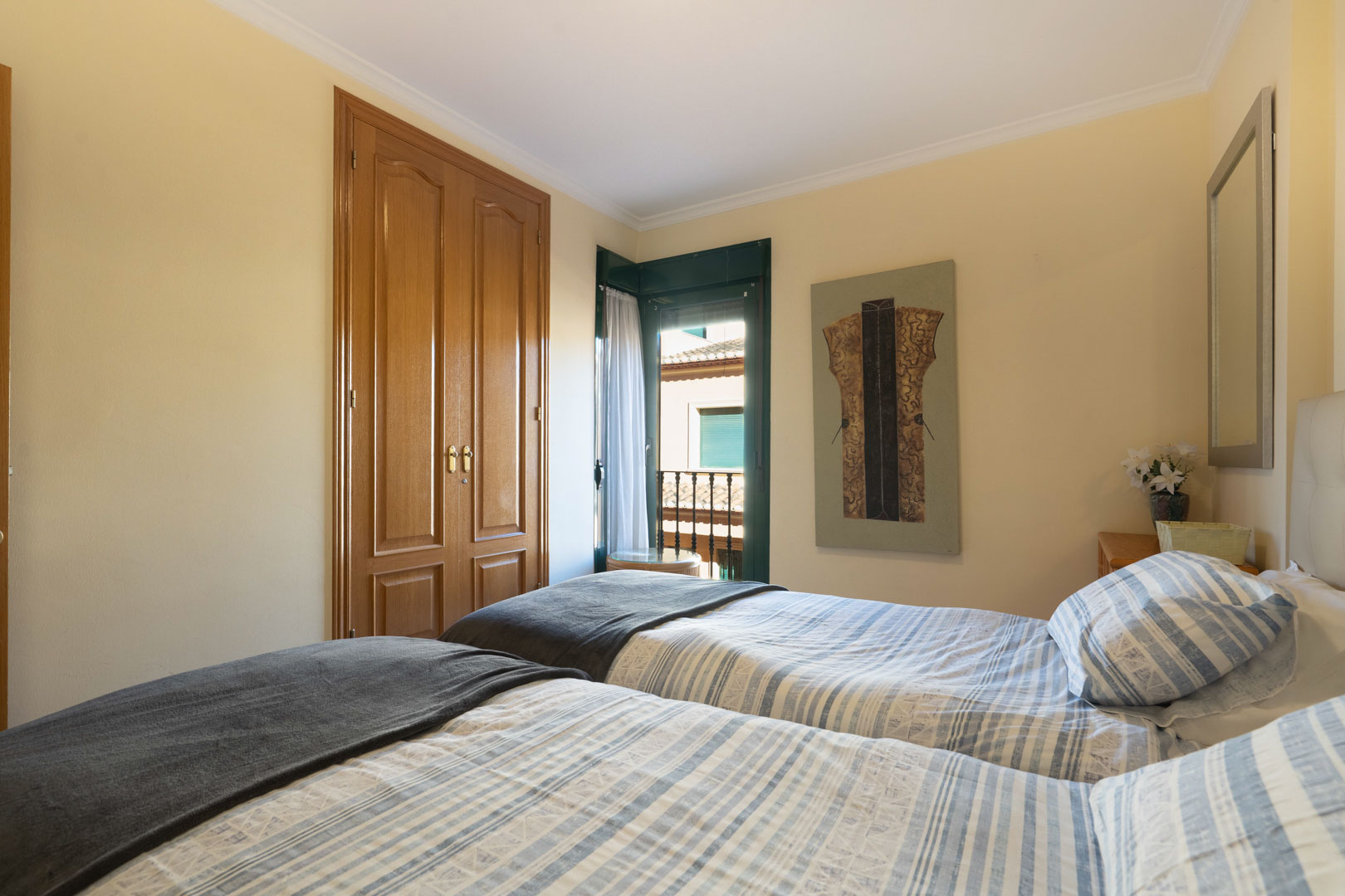 Lichte slaapkamer met twee eenpersoonsbedden, houten inbouwkasten en directe toegang tot een balkon met traditionele groene luiken.