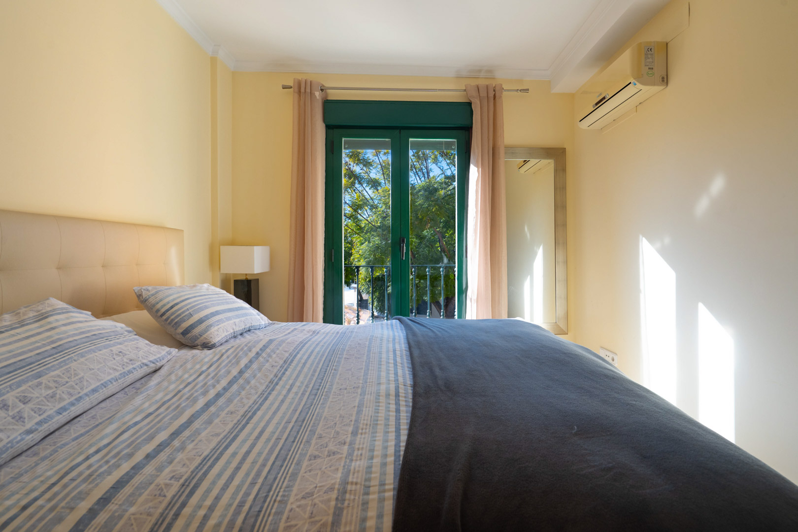 Slaapkamer met tweepersoonsbed, airconditioning en groene balkondeuren. De kamer geniet van natuurlijk licht en uitzicht op de bomen.