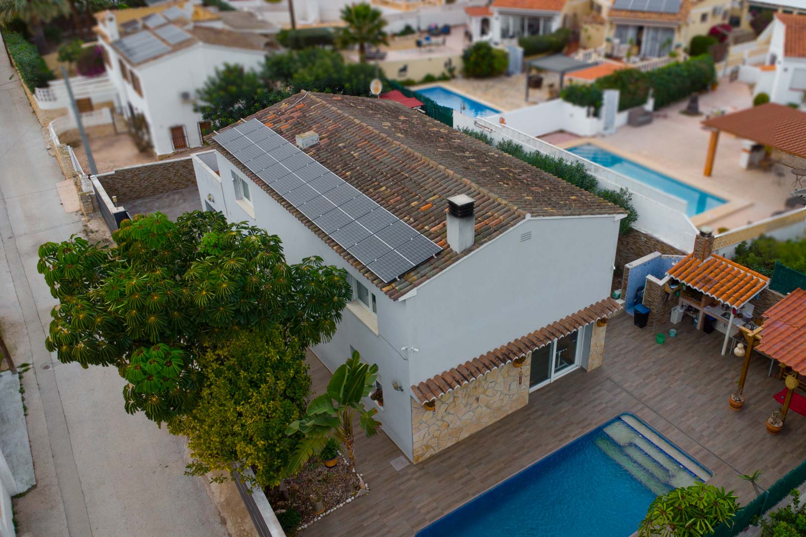 Luchtfoto van de villa met zonnepanelen op het dak, een privézwembad en een buitenkeuken op een betegeld terras omgeven door groen.