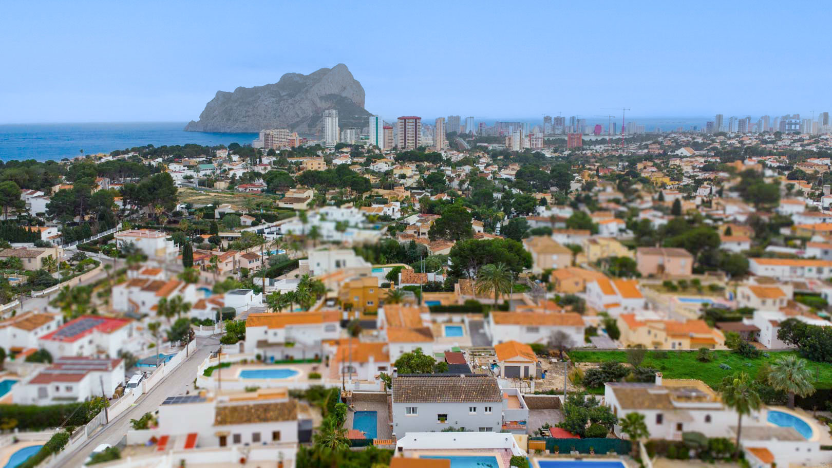 Panoramisch luchtfoto van de woonwijk Carrio in Calpe, met de iconische Peñón de Ifach en de Middellandse Zee op de achtergrond onder een heldere hemel.