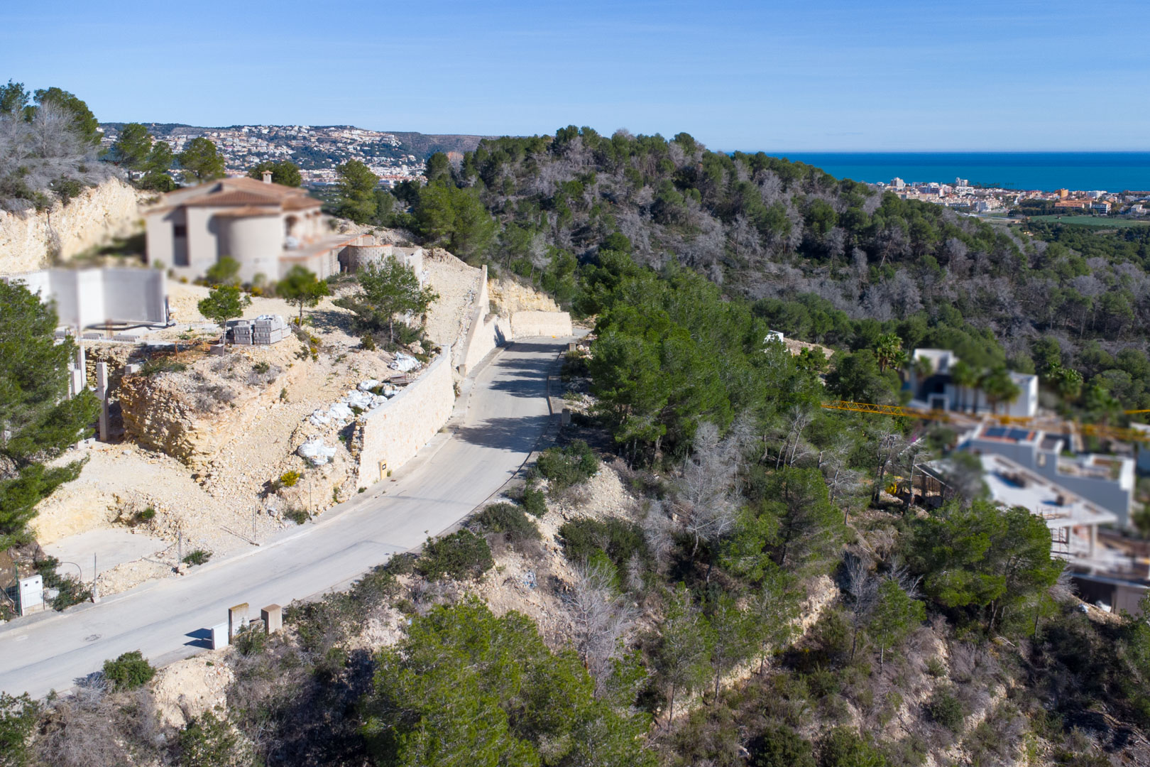 Luchtfoto van de geasfalteerde toegangsweg in Tosalet 5, met stenen keermuren, pijnbomen en uitzicht op de Middellandse Zee en de kustlijn van Jávea.