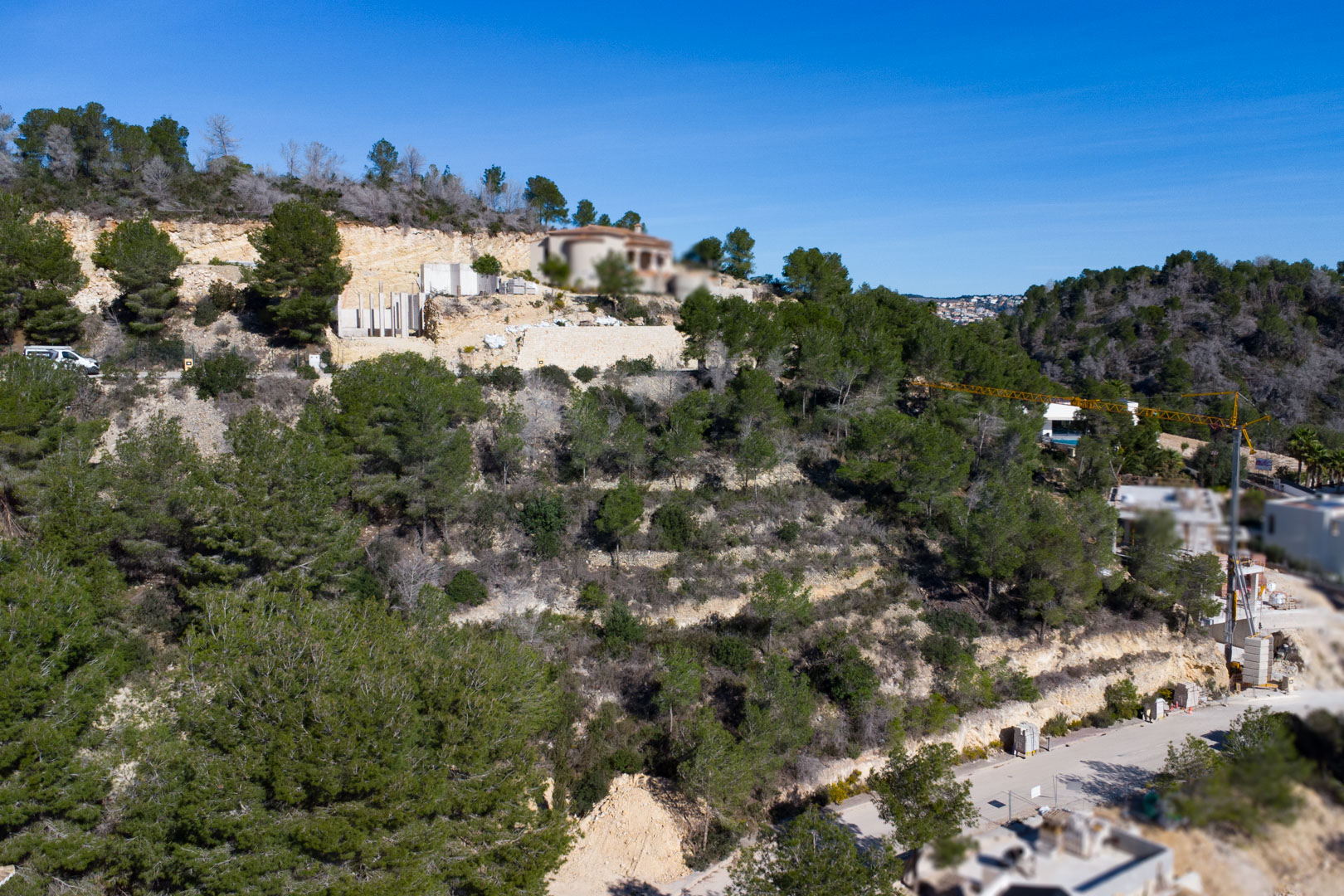 Luchtfoto van een hellend perceel met mediterrane pijnbomen, stenen keermuren en lopende bouwwerkzaamheden in de wijk Tosalet 5.