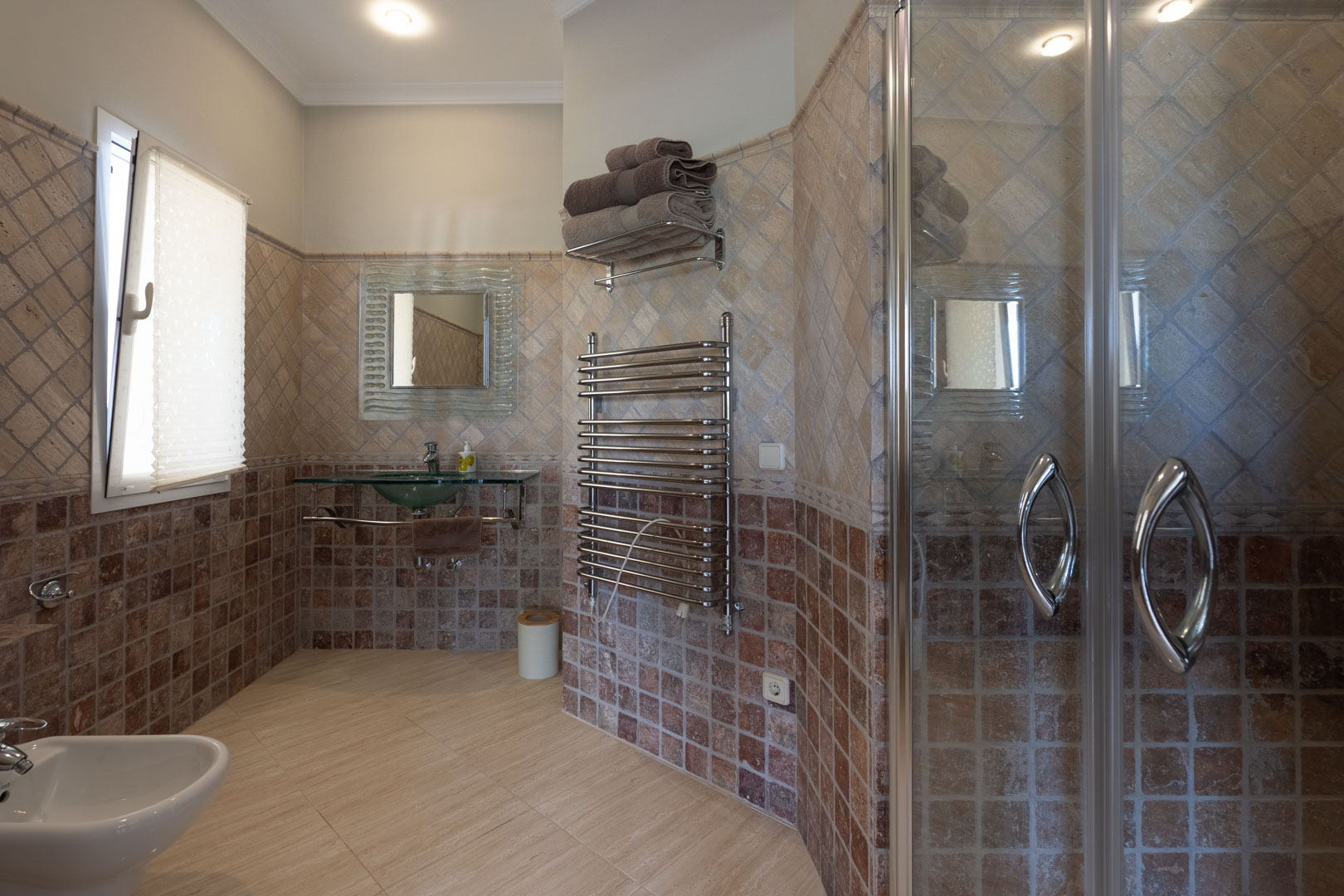 Badkamer met aardetinten wandtegels, een glazen wastafel, chromen handdoekradiator en een glazen douchecabine met inbouwspots.