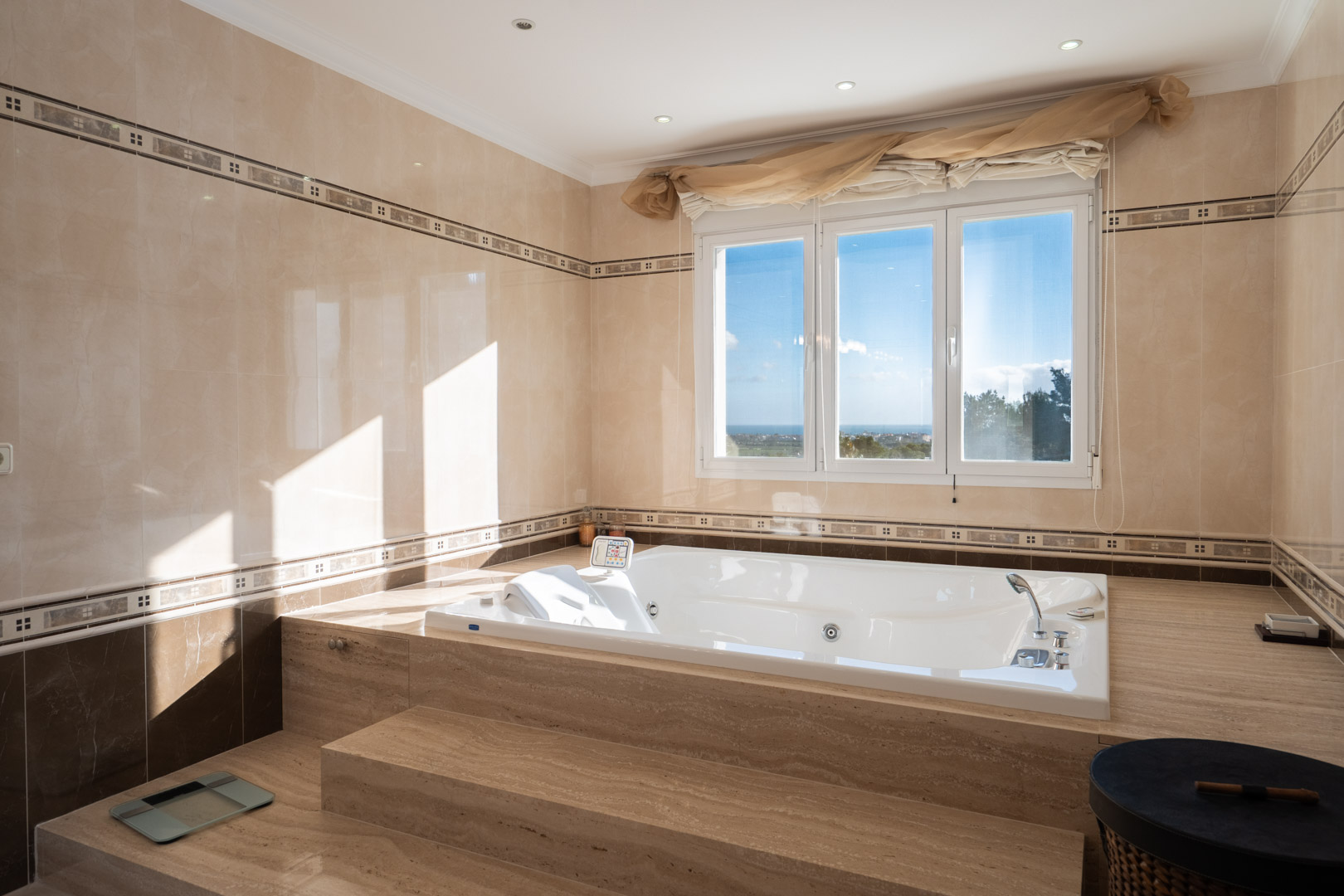 Badkamer met een in marmer ingebouwd hydromassagebad, een groot raam met uitzicht op zee en wanden afgewerkt met decoratieve sierlijsten.
