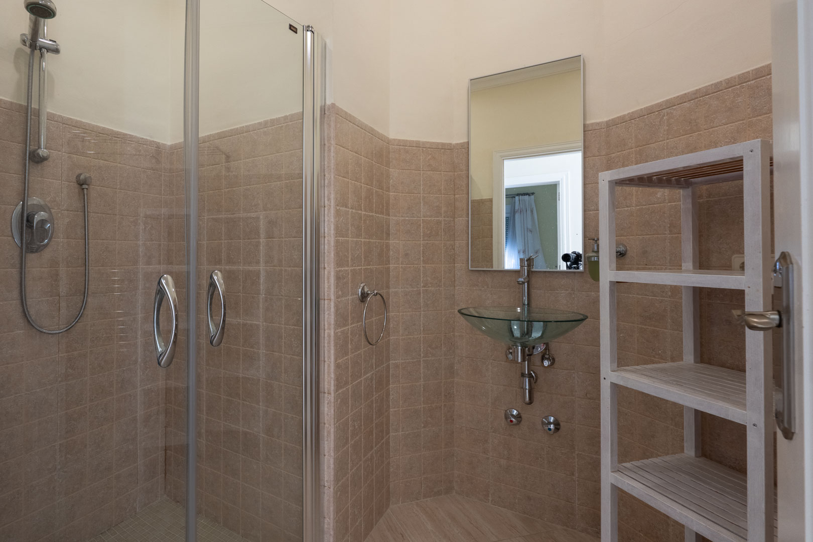 Badkamer voorzien van een glazen douchecabine, een unieke glazen waskom, beige wandtegels en een wit houten opbergrek voor handdoeken.