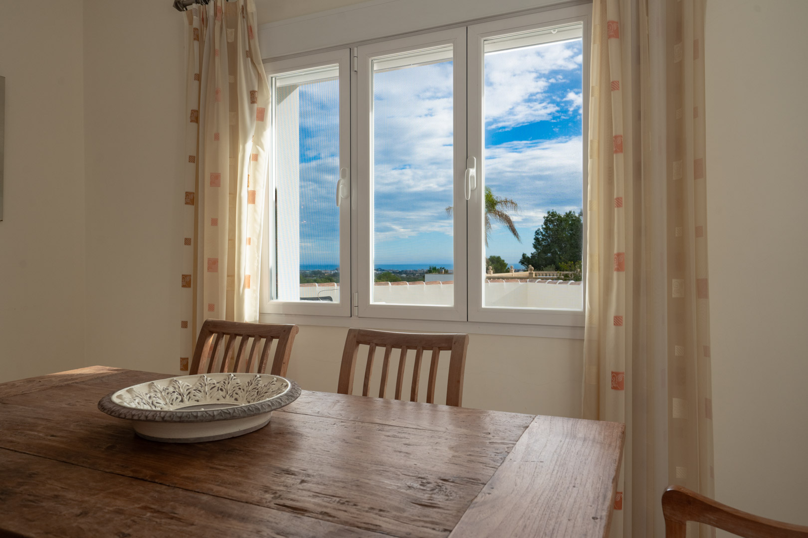 Eethoek met een robuuste houten tafel en uitzicht op de Middellandse Zee via een wit drieluiksvenster dat zorgt voor een lichte en open sfeer.