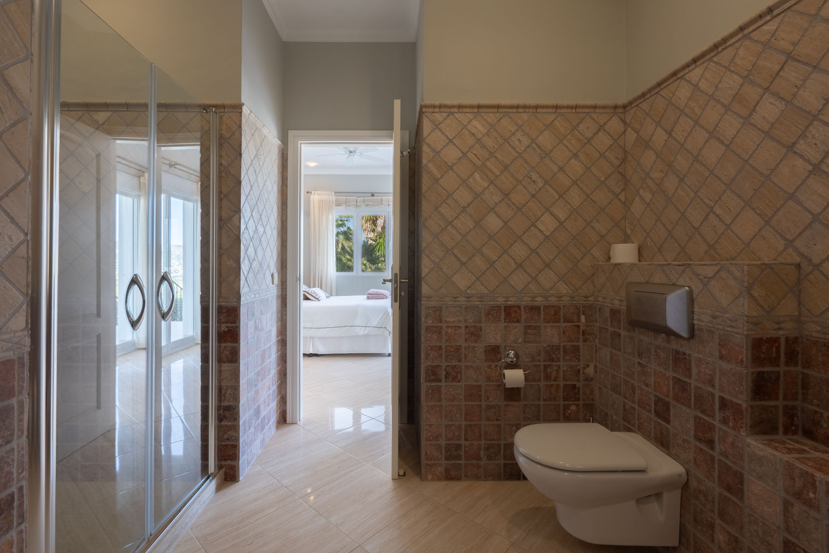 En-suite badkamer met rustieke stenen tegels, een hangend toilet en een open verbinding met de lichte hoofdslaapkamer.