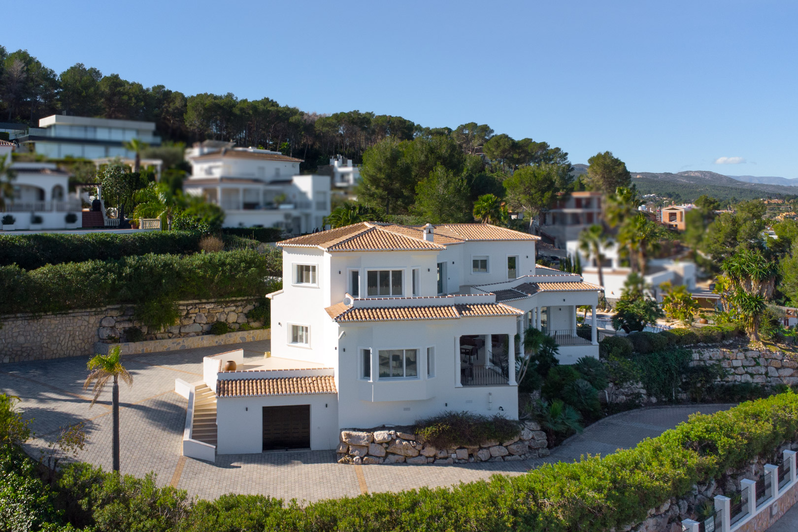 Exterieur van de witte mediterrane villa met terracotta daken, een ruime geplaveide oprit en een geïntegreerde garage op het onderste niveau.