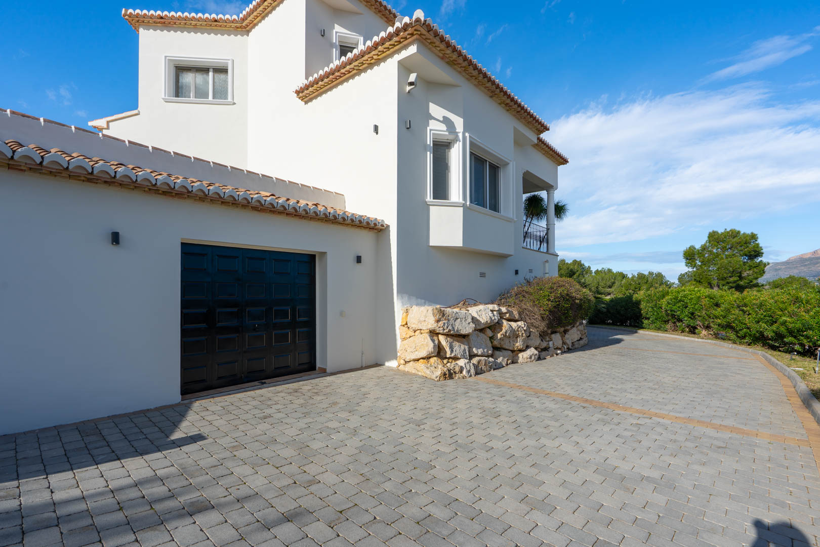 Geplaveide oprit met toegang tot de garage, gekenmerkt door witte mediterrane architectuur en robuuste natuurstenen keermuren naast de woning.