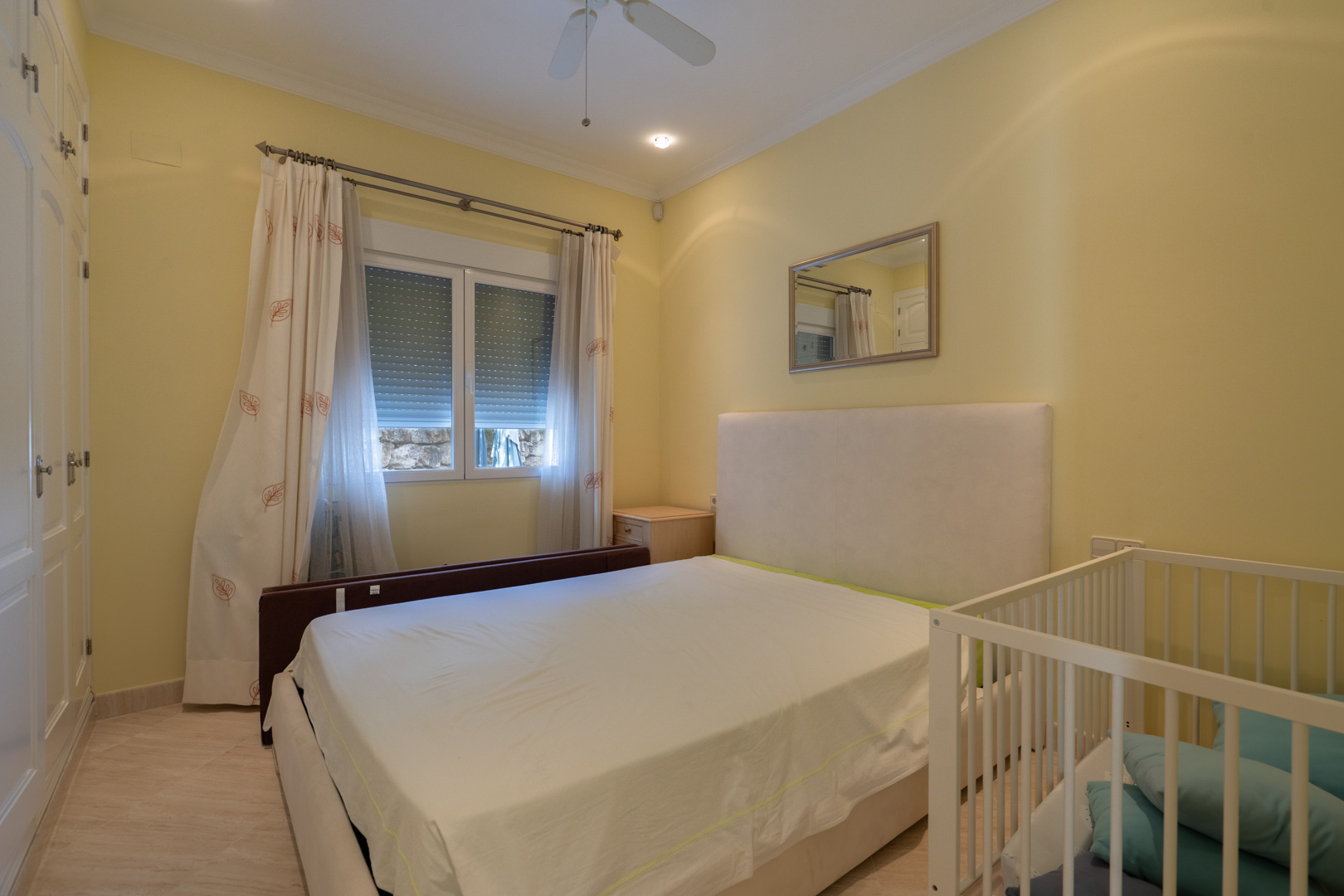 Lichte slaapkamer met crèmekleurige muren, witte inbouwkasten, een tweepersoonsbed met gestoffeerd hoofdbord en een babybedje. Voorzien van een plafondventilator en inbouwspots.
