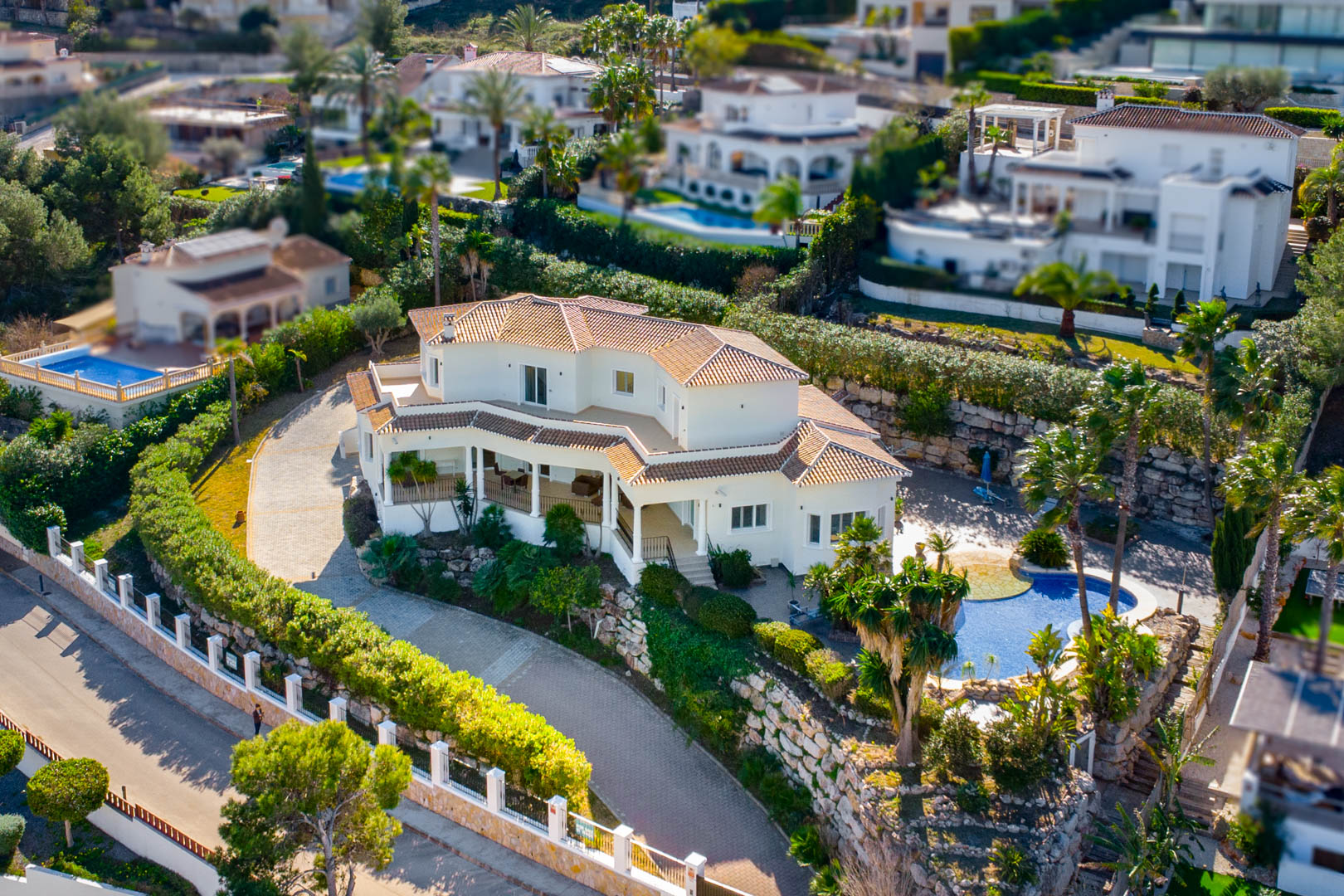 Luchtfoto van de villa met witte gevels en pannendak, voorzien van een bestrate oprit, een privézwembad en weelderige mediterrane tuinen op een dubbel perceel.