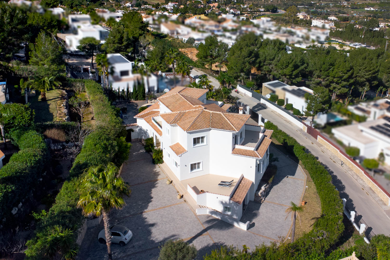 Luchtfoto van de witte villa met keramische dakpannen, een ruime geplaveide oprit en diverse privéterrassen omgeven door mediterrane begroeiing.