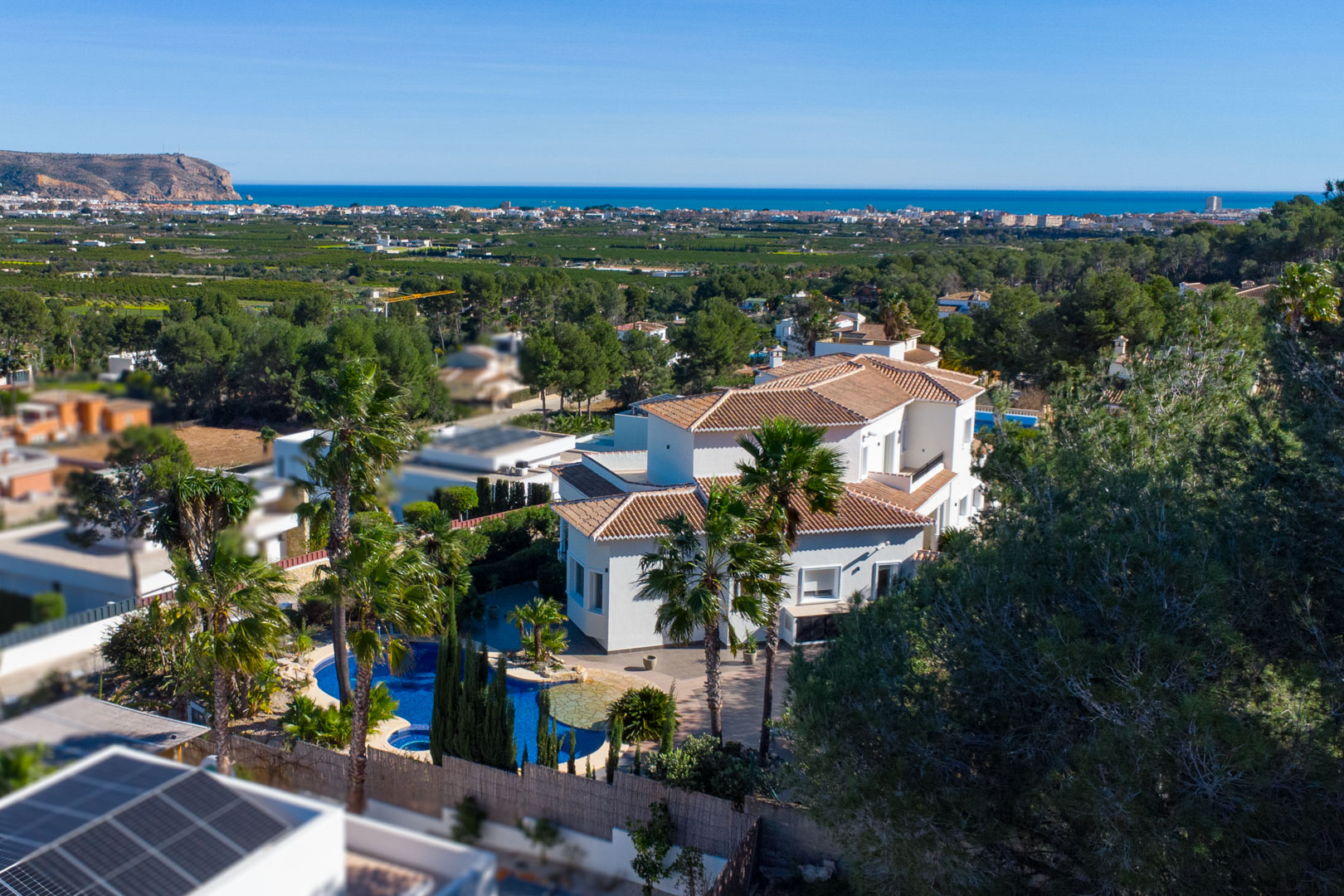 Luchtfoto van de witte villa met pannendaken, voorzien van een zwembad met jacuzzi en een mediterrane tuin met weids uitzicht over de kust en de zee.