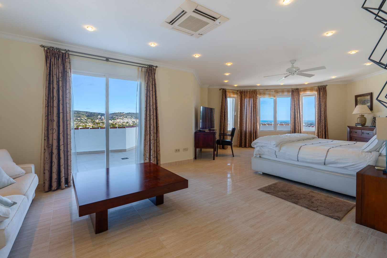 Master suite met tegelvloer, zithoek en toegang tot een privéterras met uitzicht op zee. Voorzien van klassiek houten meubilair en inbouwverlichting.