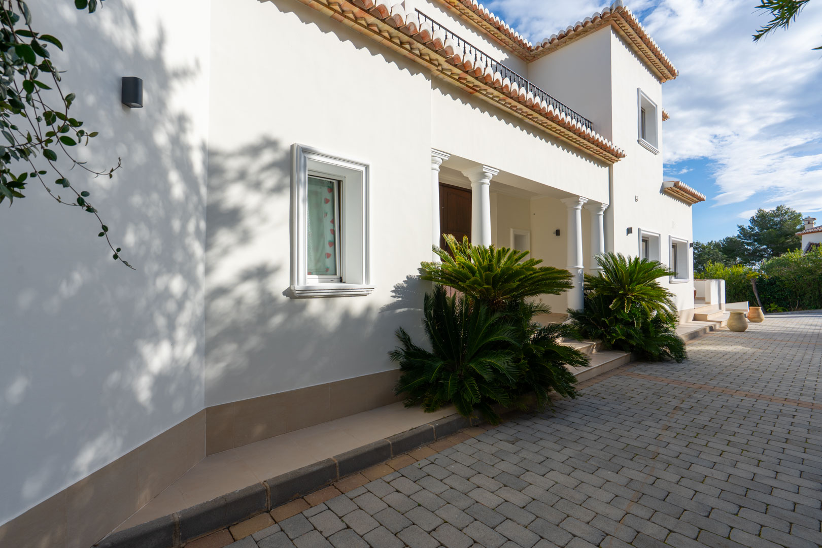 Mediterrane gevel met witte kolommen, een geplaveide oprit en architectonische details zoals terracotta dakpannen en weelderige beplanting.