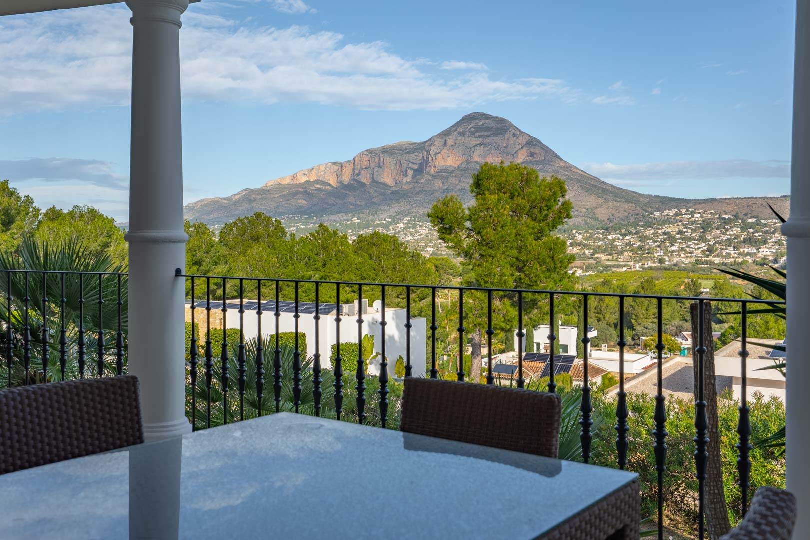 Overdekt terras met een eethoek buiten en smeedijzeren balustrade, met een panoramisch uitzicht op de berg Montgó.